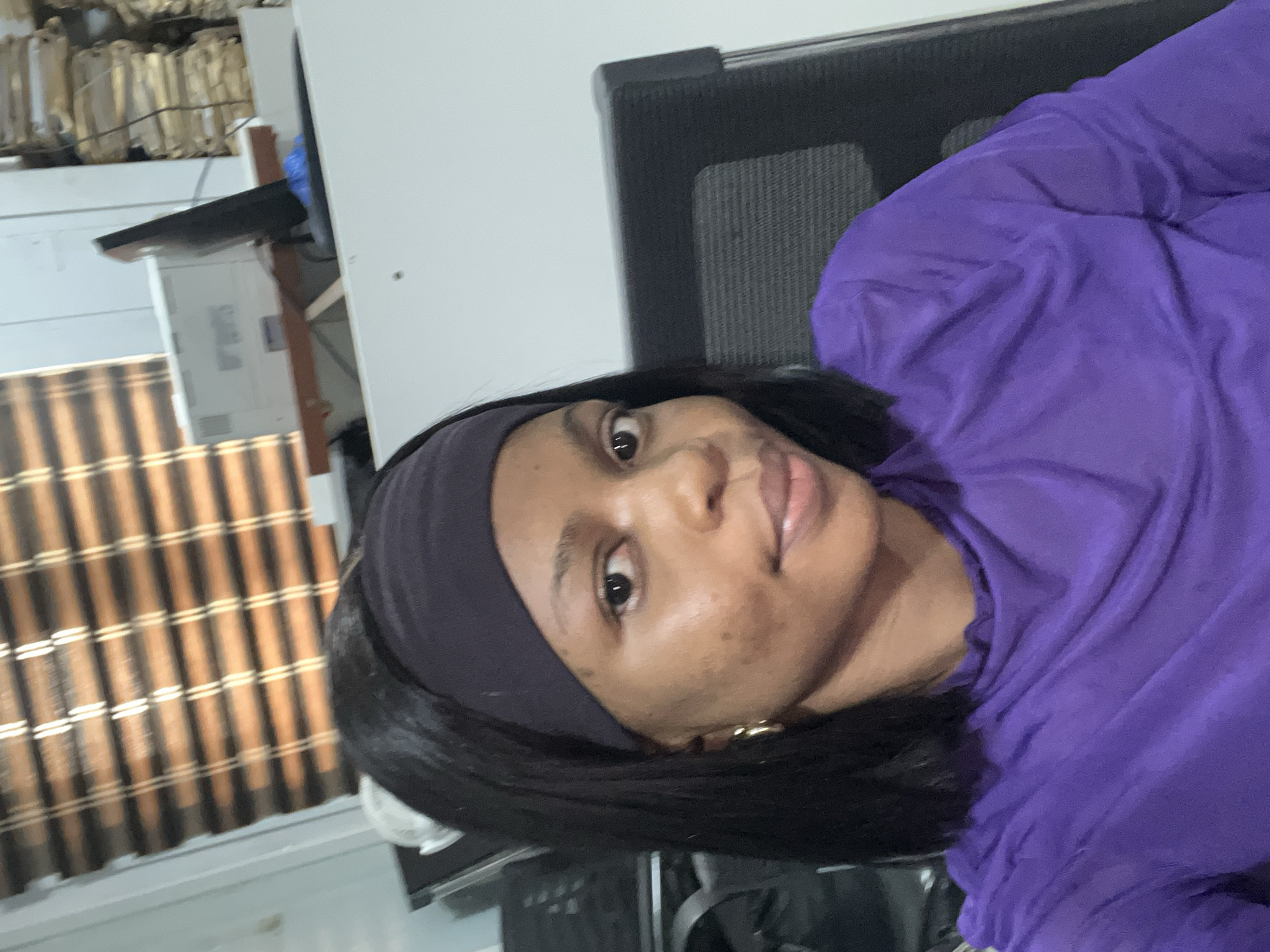A medium, color, indoor photograph depicts a Black woman wearing a black headband and a purple long-sleeved shirt. The woman is seated in a black office chair with a mesh back. Behind her, to the left, is a window covered with a brown and tan woven blind. Further back, on a white desk, are what appears to be stacked files and a monitor. The image appears to be taken indoors in the city of Ikeja, Nigeria.