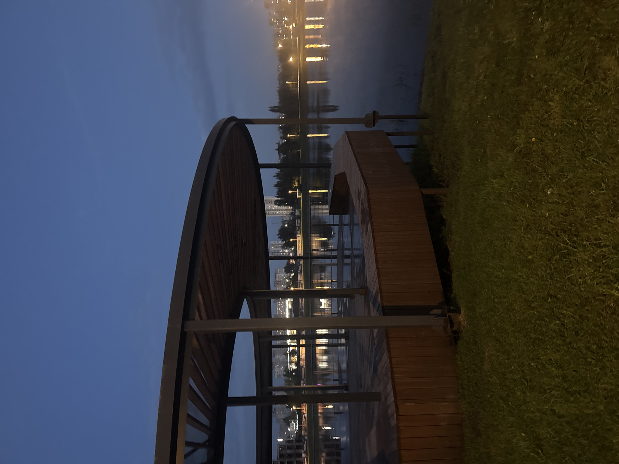 The image depicts a circular wooden pavilion with a metal frame situated next to a calm body of water at dusk. The pavilion has a wooden deck and a wooden bench. Reflections of city lights are visible on the water's surface, indicating an urban setting. In the background, there are multi-story buildings and trees. The sky is a deep blue, suggesting the time is either early morning or late evening. The ground in the foreground is covered with green grass. This scene is characteristic of a waterfront park or recreational area in the city of Ufa, Russia, possibly in a newly developed area like the Ufa River embankment or a park with water features, given the modern architecture of the pavilion and the presence of apartment buildings.