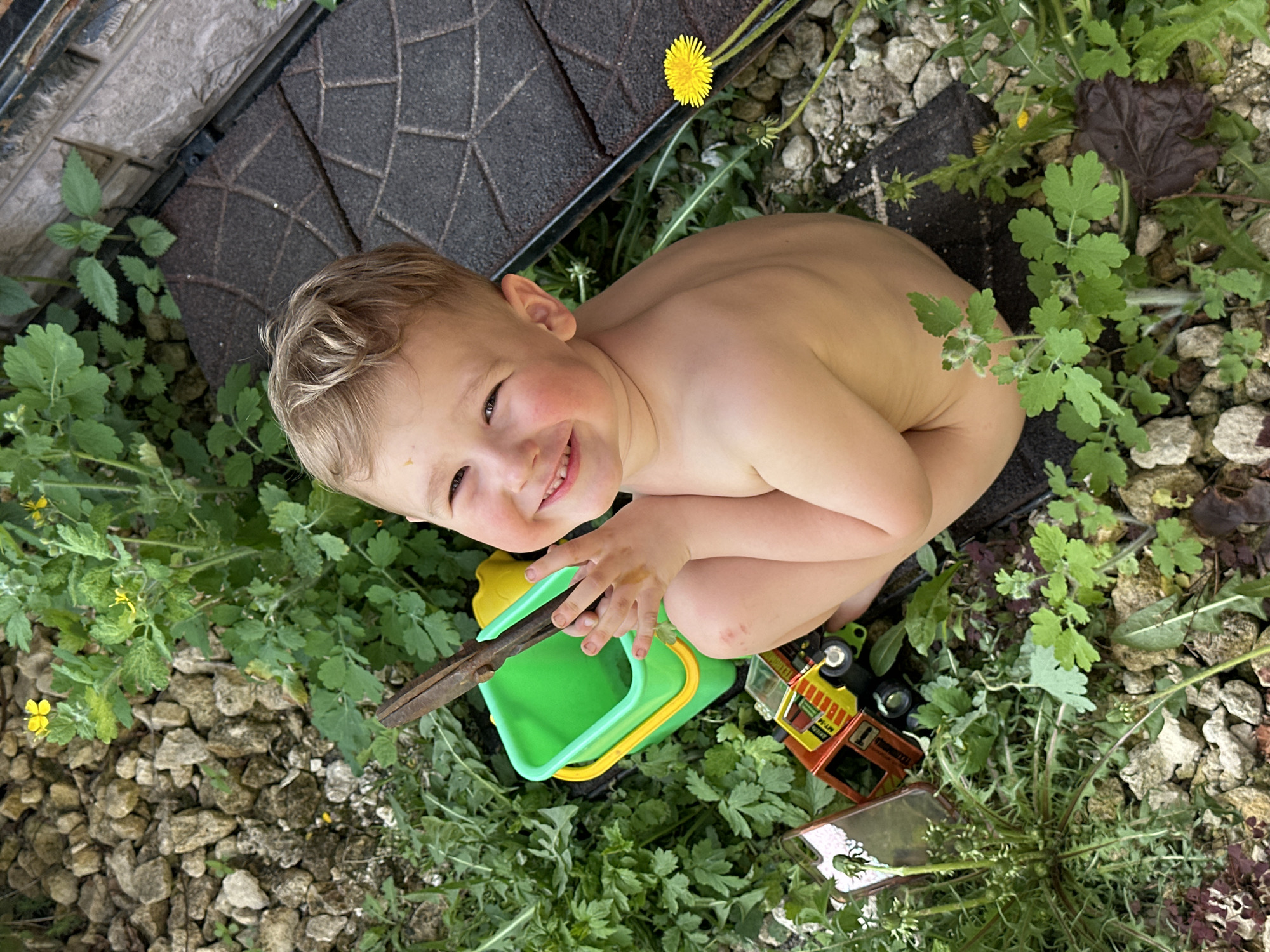 A child with blonde hair is sitting amidst rocks and foliage, near a section of brown, flat tiles. The child is shirtless, smiling, and facing the camera. A green and yellow plastic bucket containing a stick, and a small toy truck are nearby. A dandelion flower is visible. The location is an outdoor space. There is no evidence to confirm that the image was taken in Nizhny Novgorod, Russia.