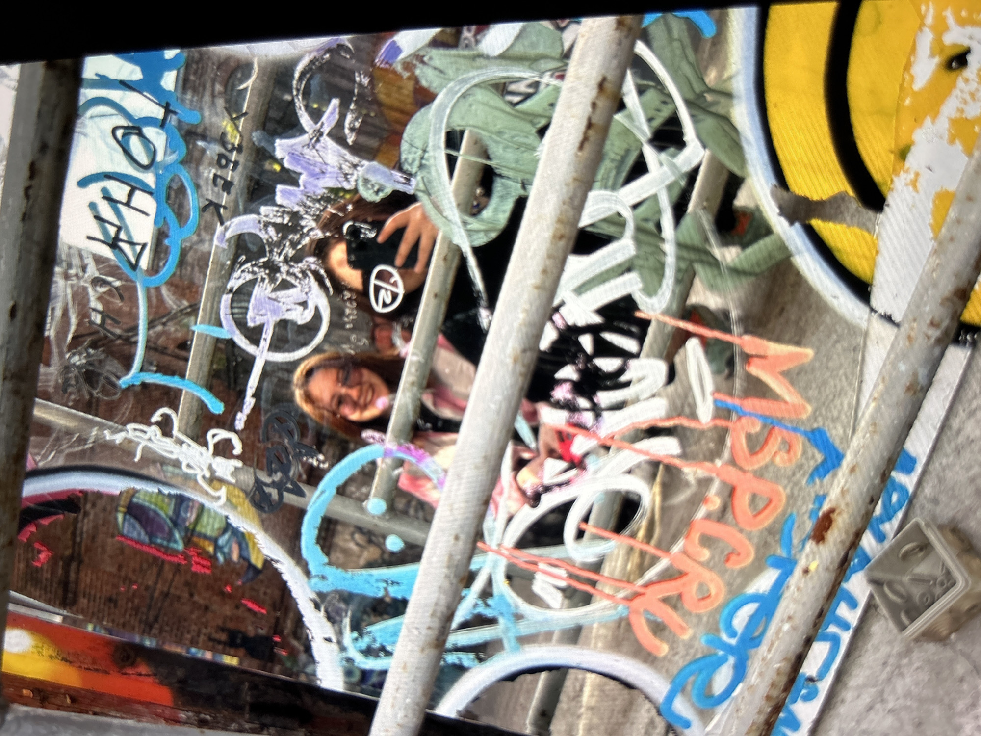 The image is a close-up, slightly blurred, vertical shot of a reflective surface covered in graffiti. Two young women are visible in the reflection. The woman on the left is wearing a pink and white patterned jacket and glasses, and is smiling at the camera. The woman on the right is wearing a black hoodie and is holding up a black smartphone to take a selfie. The graffiti is colorful and abstract, with various tags and images in blue, pink, white, and green. The surface appears to be glass or a similar material, as it reflects the surrounding environment, including a brick wall and what looks like metal bars. The overall impression is of an urban, gritty setting, likely in a city. In the lower foreground, there are metal bars and a small plastic container filled with screws or washers. The image is taken from a low angle, looking up towards the graffiti and the women.