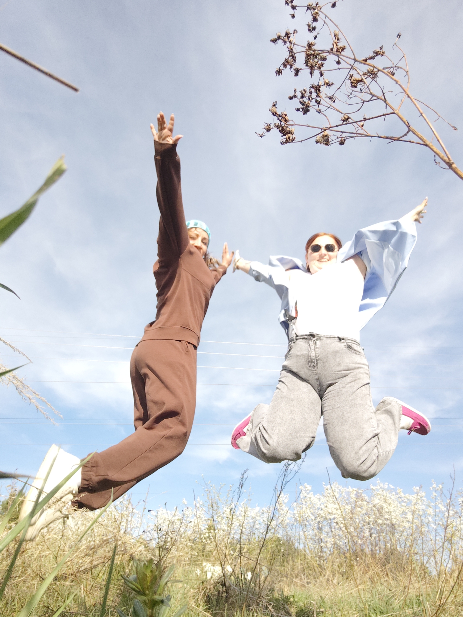 Two young women are jumping in the air. One woman is wearing a brown sweatsuit and a blue headband. The other woman is wearing a white shirt, grey jeans, a light blue jacket, and pink shoes. They are in a grassy field with white flowers and a blue sky in the background. A leafless branch is in the upper right corner of the image. The location is Chaikovsky, Russia.