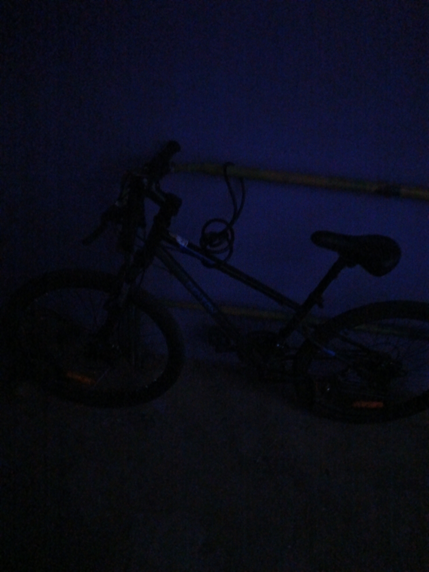 In an indoor environment, a dark bicycle with a black frame is the primary subject, positioned in front of a dark blue wall. A yellow pipe runs horizontally across the upper portion of the frame, behind the bicycle's handlebars. The bicycle has two wheels, both black, with visible spokes. The front wheel is on the left and the rear wheel is on the right. A black seat is mounted on a black seat post. The bicycle appears to be locked with a coiled black cable lock wrapped around the frame. The lighting is dim, casting a shadow over much of the scene. No people are present in the image.
