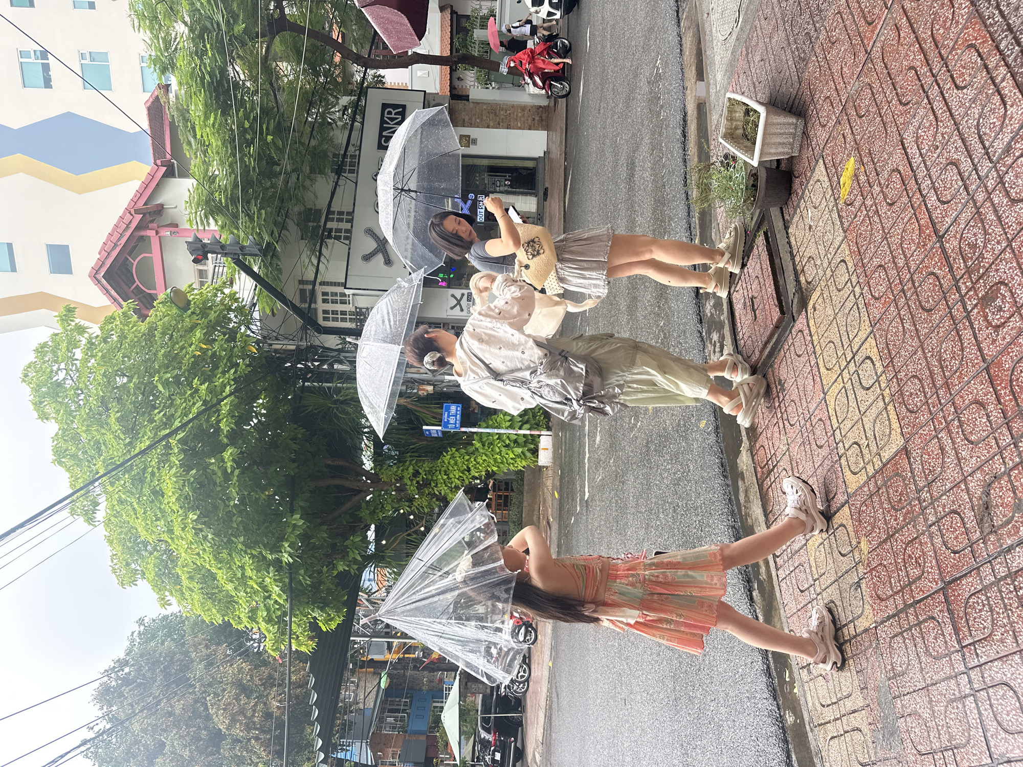Three women are standing on a wet tiled sidewalk in Nha Trang Ward, Vietnam, using umbrellas in rainy conditions. The woman closest to the camera is wearing a strapless dress and holding a clear umbrella over her head. The second woman, slightly further back, is wearing light-colored pants and a white top, also holding an umbrella. The third woman, furthest away, wears a short striped dress and is holding a bag.
Behind them are trees, overhead electrical wires, and buildings. One of the buildings has "CNKR" written on its facade. Vehicles, including scooters and cars, are visible on the street. A street sign is also visible. A planter sits on the sidewalk.