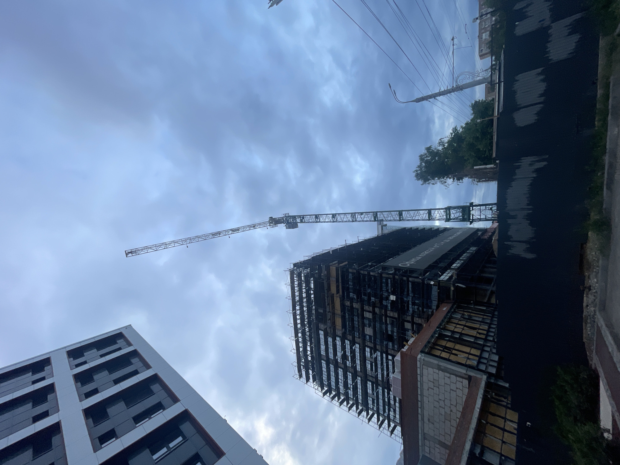 This image captures an urban construction site in the city of Unknown, Unknown, viewed from a low-angle perspective under an overcast, grey-blue sky.
In the immediate foreground, a dark, corrugated metal fence, exhibiting vertical lighter streaks, acts as a barrier, with a narrow strip of ground and a concrete curb visible beneath it. To the far left, a completed, modern high-rise building with a light-colored facade and dark, rectangular windows is partially visible.
Centrally positioned, a multi-story building is under active construction, heavily enveloped in metal scaffolding. This structure features dark exterior cladding, and a large white sign on its side displays Cyrillic text, which translates to "Class A and B Offices." Directly below this building, a smaller, lower structure with light-colored cinder block walls and a reddish-brown sloped roof is also under construction, revealing exposed framework and yellow paneling.
To the right of the central construction, a tall, grey lattice tower crane stands prominently. Further to the right, a utility pole supports multiple overhead power lines that traverse the frame. Scattered trees and vegetation are visible behind the fence, with the faint outlines of additional buildings in the distant background. No individuals are discernible within the frame.