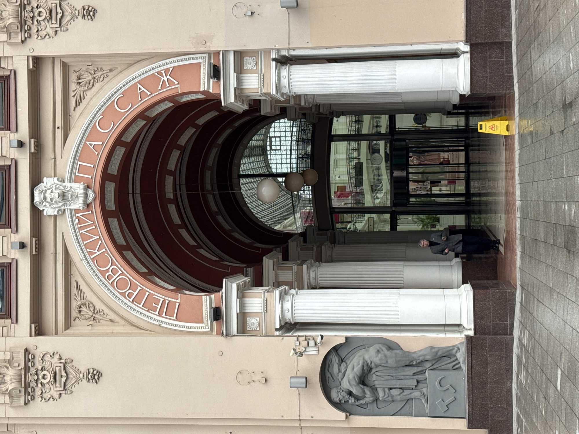 The image captures the ornate entrance to the Petrovsky Passage in Moscow, Russia, viewed from the exterior looking inward.
The central focus is a grand archway, adorned with intricate architectural details. Along the curve of the arch, prominent white Cyrillic text reads "ПЕТРОВСКИЙ" (Petrovsky) on the left and "ПАССАЖ" (Passage) on the right, identifying the location. The inner surface of the arch is a reddish-brown with white decorative panels, while the surrounding facade is a light beige. At the apex of the arch, a detailed lion's head carving serves as a keystone. Further ornate carvings, resembling floral motifs and classical patterns, decorate the spandrels above the arch.
The entrance is flanked by sturdy, fluted white columns with elaborate capitals, set against the beige facade. Below the right-hand columns, a large, dark grey relief sculpture depicting classical figures, including a reclining male figure, is integrated into the building's base.
Beyond the archway, the interior of the passage is visible, featuring a high, possibly glass-paneled ceiling that allows light to filter in. Several spherical light fixtures hang within the passage. Reflections of shop windows or other interior elements are visible in the glass and polished surfaces inside.
On the paved walkway leading into the passage, a single person, dressed in a dark jacket and trousers, stands inside the entrance towards the right. A bright yellow "wet floor" sign is placed on the ground just inside the entrance, near the person. The ground outside the entrance consists of dark grey and brown paving stones. The scene conveys a sense of historical elegance and grandeur, likely during daytime given the general illumination.