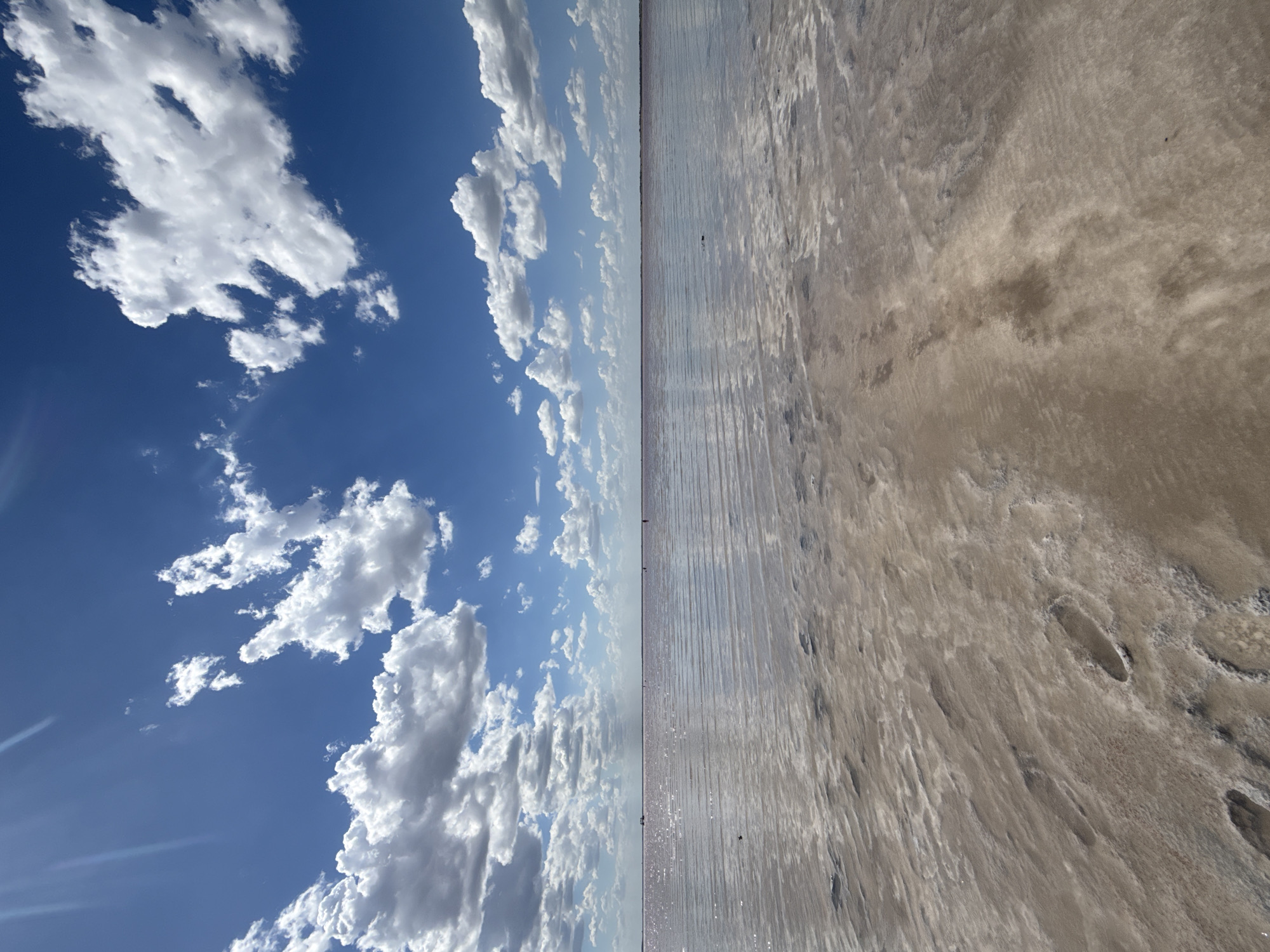 The image depicts a vast, shallow body of water, likely a salt lake, under a bright blue sky dotted with fluffy white clouds. The water exhibits a subtle pinkish hue, suggesting high salinity. The foreground shows a textured, sandy or muddy shoreline with visible footprints and ripples, indicating the shallow nature of the water. In the distance, several small figures can be seen wading or standing in the water. The horizon line is indistinct, merging the water and sky. The overall impression is one of a serene, natural landscape under a clear, sunny day. The location is identified as Bursol', Russia.