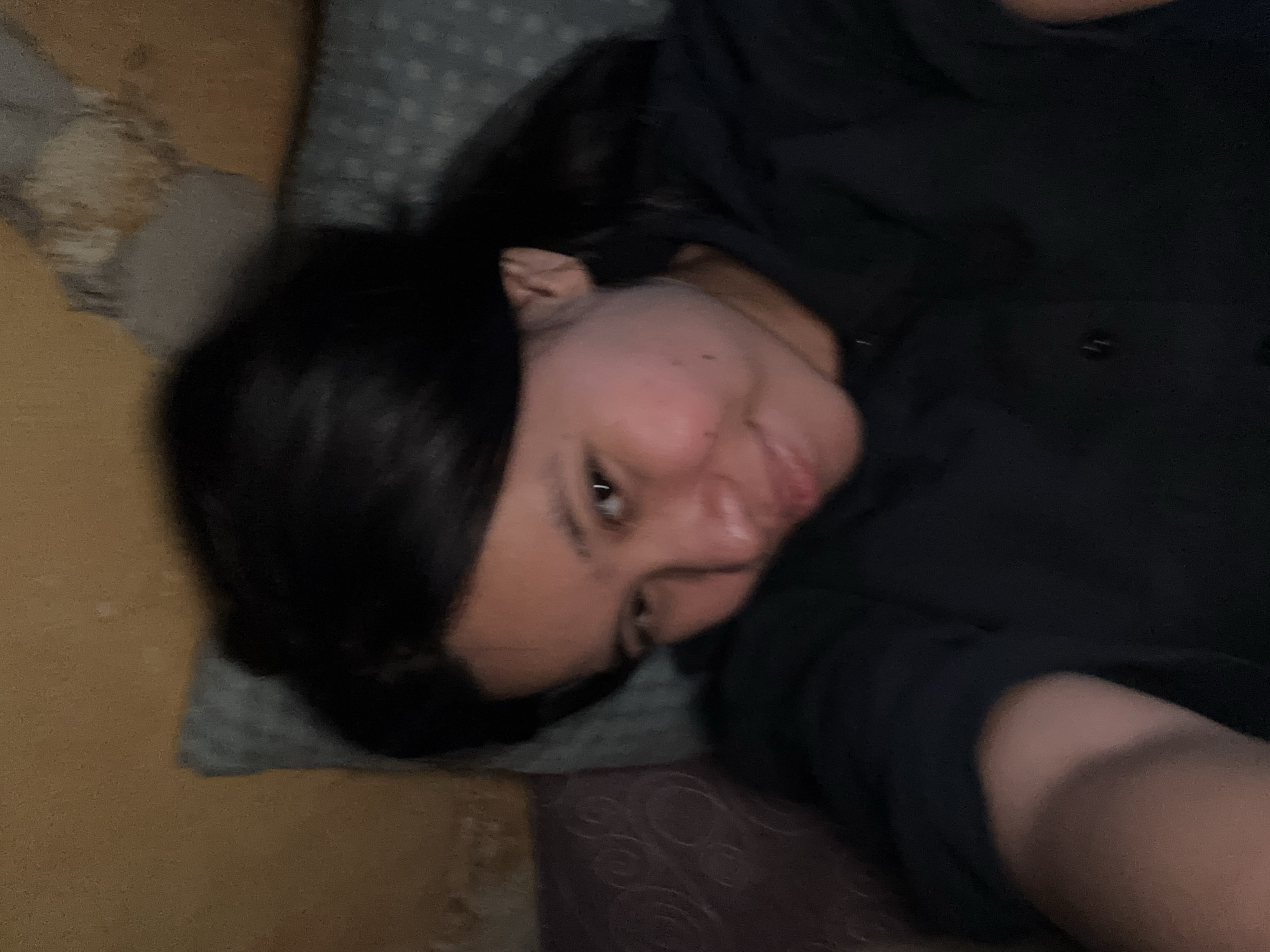 A young woman with black hair is lying down, looking at the camera. She is wearing a black collared shirt. The background appears to be a wall and a patterned pillow. The lighting is dim, suggesting the photo was taken indoors. The image is slightly blurry, indicating movement during capture. The location is not discernible from the visual information provided.