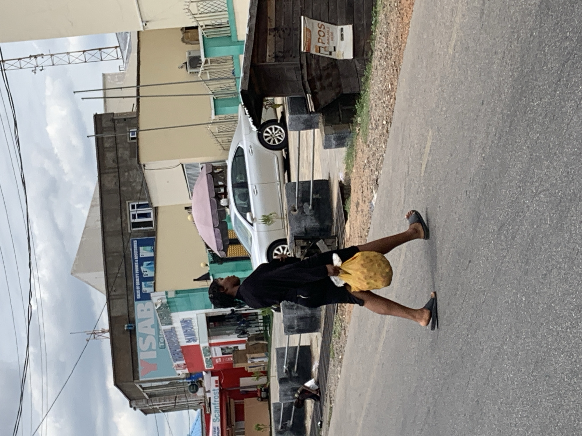 A person is walking on a street in Lokoja, Nigeria. The person is wearing dark clothing and flip-flops and carrying a yellow bag. The street is paved and there are buildings on either side of the street. The building on the left side of the image has a sign that says "VISAB". There is a white car parked in front of the building. There is a structure to the right of the building that has a sign that reads "POS MOBILE". There is a tall antenna or structure in the background on the left side of the image. The sky is overcast.