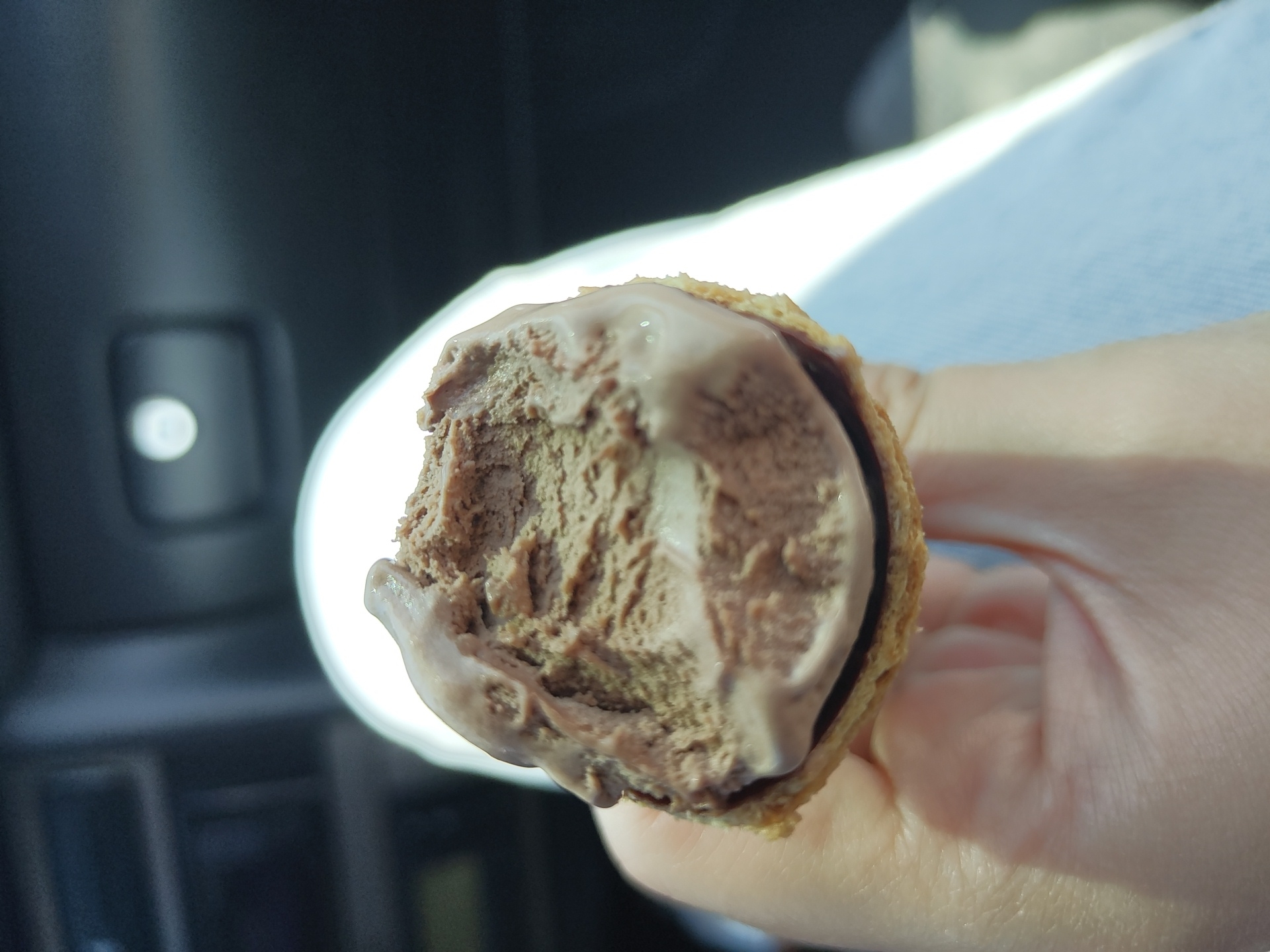 In Banichan, Bulgaria, a hand holds a chocolate ice cream cone, with the top portion of the ice cream partially eaten. The cone appears to be wafer-based. The background is blurred and appears to be the interior of a vehicle, possibly a car, with dark upholstery and a glimpse of a bright light source. The overall lighting suggests it is daytime.