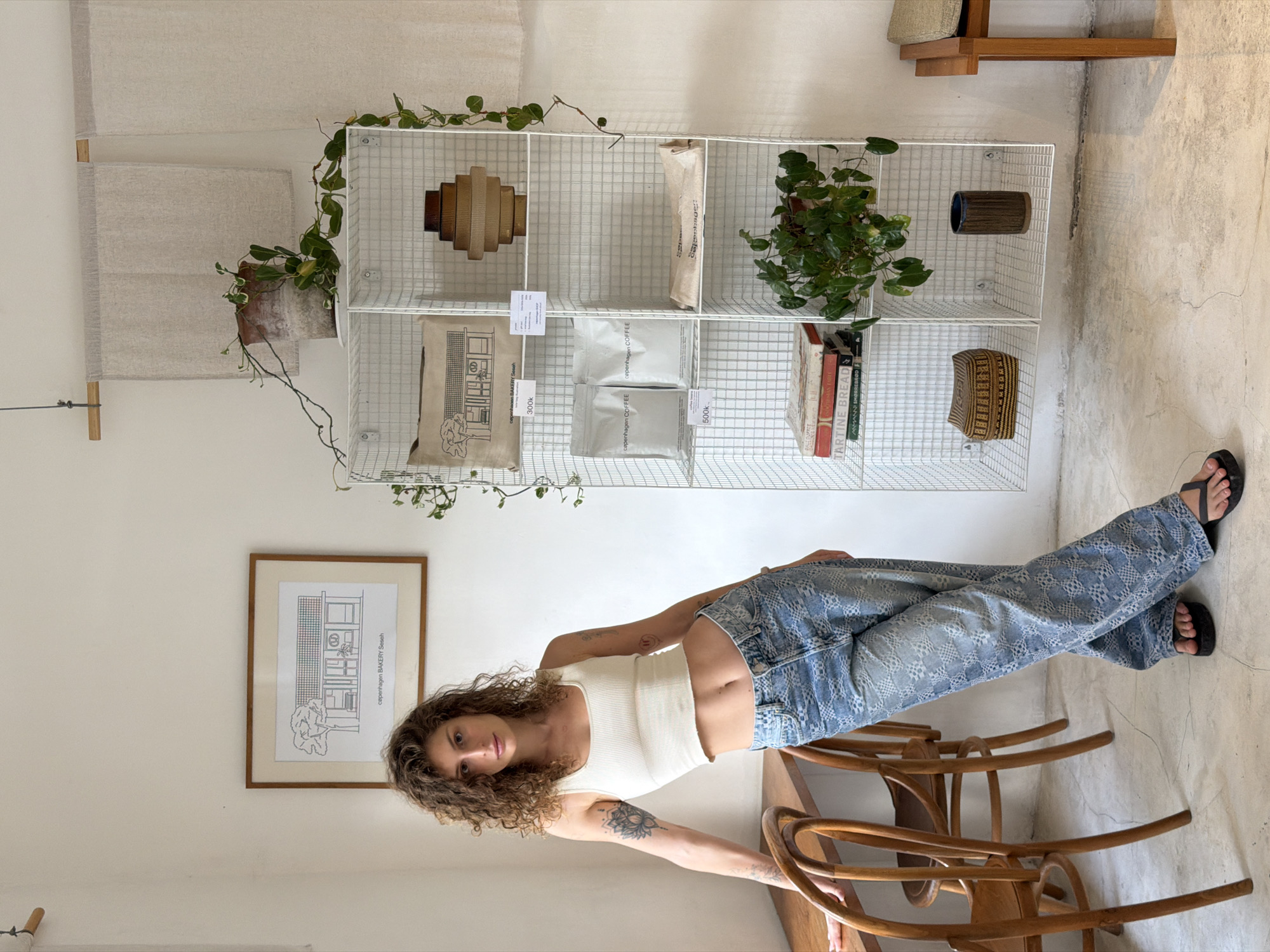 In the city of Mengwi, Indonesia, a young woman with curly brown hair stands leaning against a wooden table and chairs. She is wearing a white cropped tank top and light blue patterned jeans. Her left arm is resting on the back of a chair, and her right hand is on her hip. She is facing the camera, with a slight smile.
Behind her, a white wire shelving unit displays various items. On the top shelf, a framed drawing of a building is hung on the wall. The shelving unit also holds two bags of coffee, a stack of books, a potted plant, and decorative objects, including a brown vase and a woven basket. A vine with green leaves drapes down from the top of the unit.
The room has a minimalist aesthetic with white walls and a light-colored floor. Natural light enters from the left side of the image, illuminating the scene. A wooden chair and part of a table are visible on the left, and a wooden stool is on the right. Two light-colored cloths hang on the wall above the shelving unit. The overall impression is that of a well-lit, tastefully decorated space, possibly a cafe or a boutique.