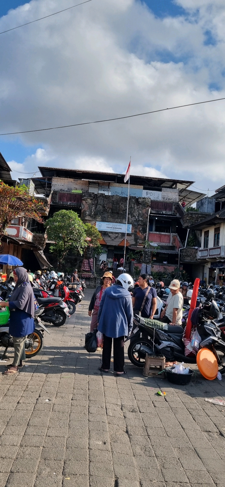 The image depicts a street scene in Denpasar, Indonesia, likely a market area based on the presence of various stalls and people. In the foreground, a paved area is filled with parked motorcycles, indicating a common mode of transportation. Several individuals are visible, some engaged in conversation, while others appear to be shopping or preparing to leave. A woman in a blue headscarf and matching tunic stands near a green cooler box and a motorcycle. Behind her, a group of people is gathered, including a woman in a hat and a blue jacket, and a man in a white shirt and a hat. The background reveals a multi-story building with a traditional stone facade and a modern roof structure, suggesting a blend of old and new architecture. A flagpole with the Indonesian flag is prominent on the building. The overall atmosphere appears to be bustling, with a mix of daily activities and commerce taking place under a partially cloudy sky.