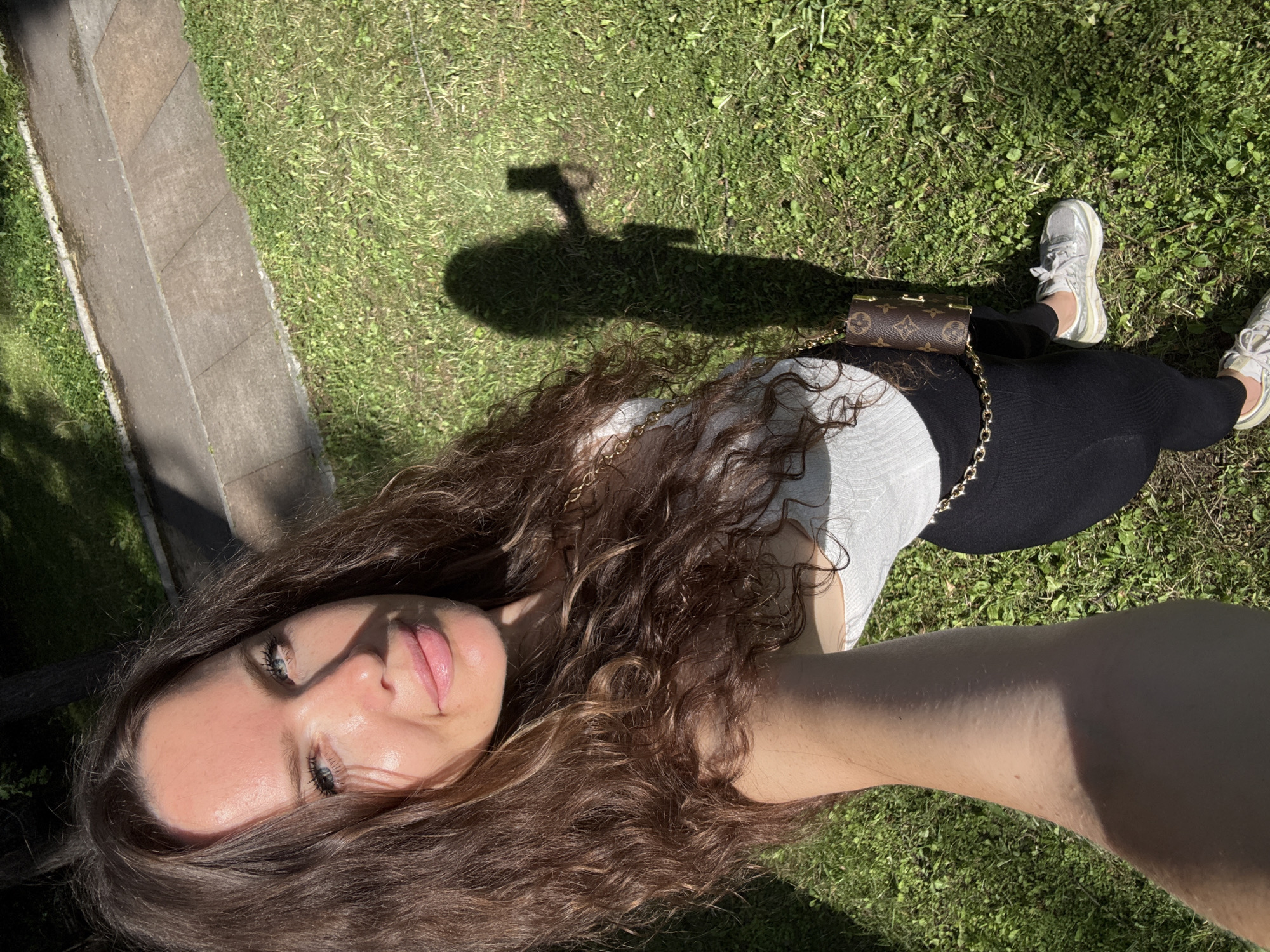 A woman is taking a selfie outdoors on a sunny day, standing on a patch of green grass. She is wearing a white tank top, black leggings, and silver sneakers. A small designer crossbody bag with a chain strap is visible. Her long, dark, wavy hair is down. Her shadow, cast by the sun, shows her holding a phone. In the background, a paved pathway runs alongside the grass. The lighting suggests it is likely a warm day.