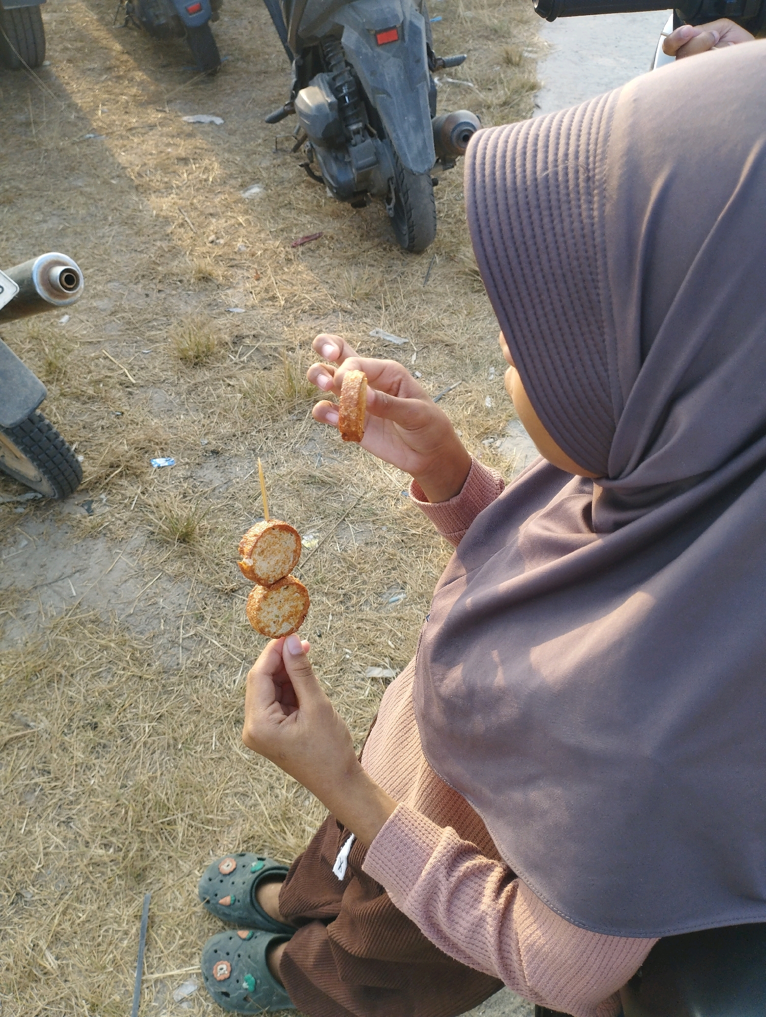 A person wearing a brown hijab and a pink shirt holds a skewer with two round, golden-brown pieces of food. The person is also wearing brown pants and green clogs with cartoon designs. The background is dry, yellowish grass with some scattered debris. In the background, there are parts of several motorcycles. The scene appears to be an outdoor area, possibly a roadside or parking lot, in Kuala Samboja, Indonesia, given the presence of motorcycles and the dry, grassy environment. The lighting suggests it is daytime, with sunlight casting shadows.