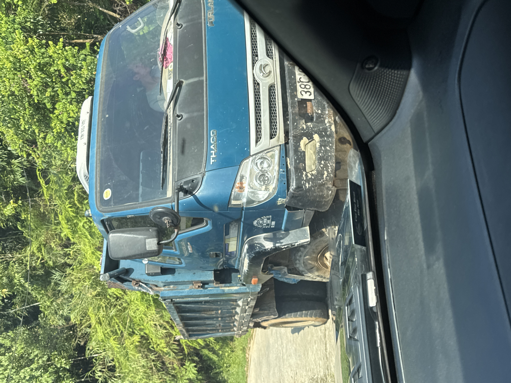 This image, taken in Thông Nguyên Commune, Vietnam, captures a blue utility truck from the perspective of a passing vehicle's interior. The photo shows the front and side of the blue truck, which appears to be a dump truck or flatbed, driving on a paved road. Dense green foliage, likely trees and bushes, lines the left side of the road, indicating a rural or semi-rural setting.
The blue truck is branded with "THACO" and "FORLAN" visible on its cab, along with a circular emblem on its grille. Its front bumper and lower sections are covered in mud, suggesting recent travel on unpaved terrain. A partial Vietnamese license plate, "38C", is visible at the front. A person's face can be faintly seen through the truck's windshield. On the side of the truck, there's a white graphic with text that appears to be Vietnamese, possibly a business name like "ĐIỆN LẠNH CÔNG TY" (Electrical/Refrigeration Company). A white air deflector is mounted on the roof of the cab, also bearing some indistinct text.
The image is captured during daylight hours, under clear and sunny weather conditions. The immediate foreground shows parts of the photographer's vehicle interior, including a speaker grille and an A-pillar trim. The scene depicts typical daily life and transport in a Vietnamese provincial area.
