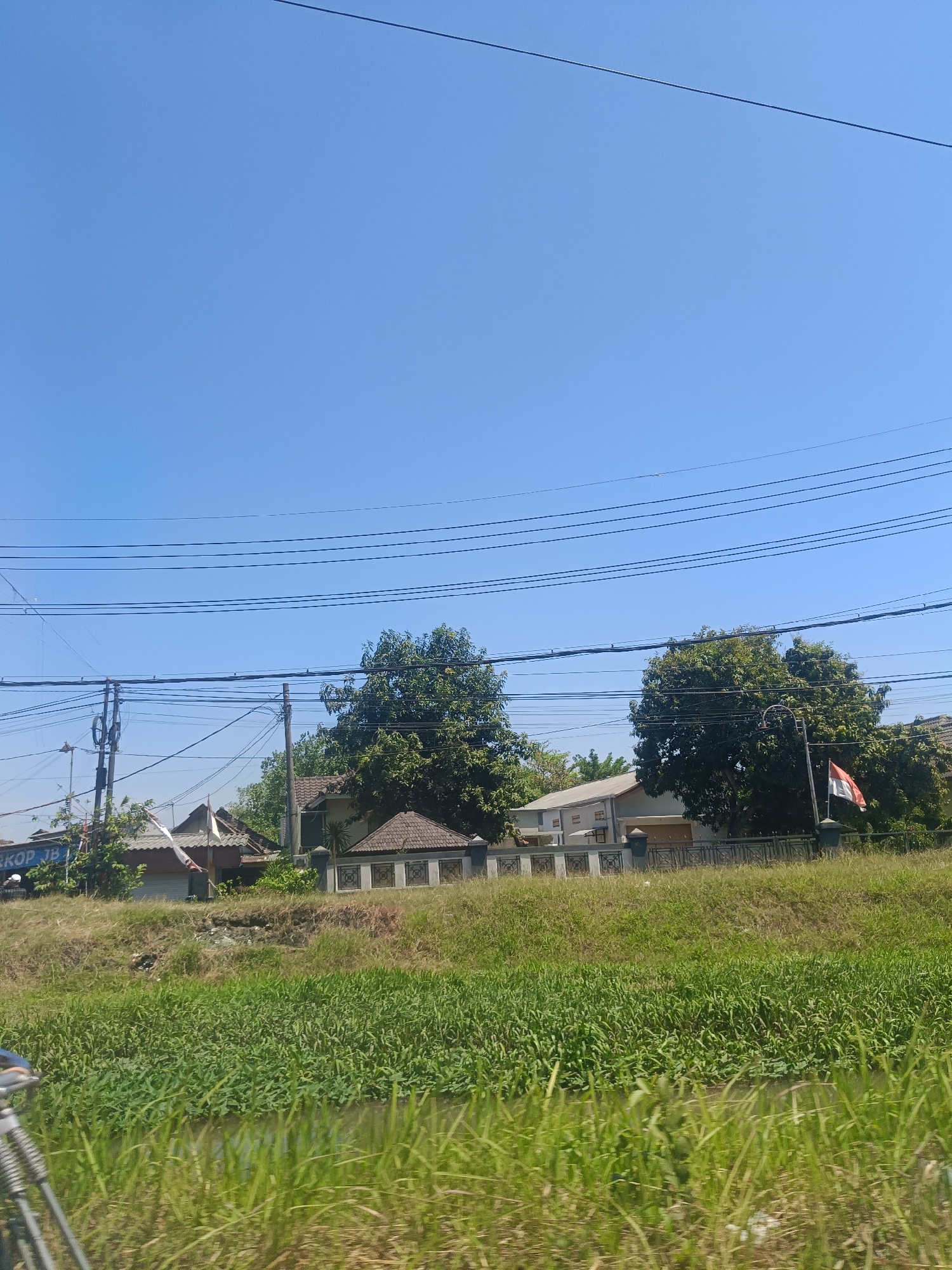 In Ganting, Indonesia, a bright, clear sky dominates the upper half of the frame, traversed by a series of black electrical wires. Below the wires, a lush green landscape unfolds, featuring a body of water overgrown with vegetation. On the slight incline above the water, several residential buildings are visible, some with tiled roofs and distinct architectural features. Trees are interspersed among the houses, adding to the verdant scenery. A flag, red and white, is mounted on a pole near one of the houses, adding a touch of color to the scene. The foreground is filled with tall, green grass, suggesting an outdoor, possibly rural or suburban, setting. The overall impression is one of a peaceful, sunlit day in an Indonesian community.