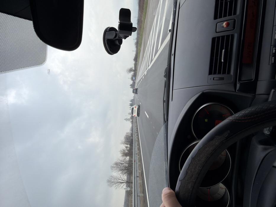 The image depicts a driver's perspective from inside a vehicle traveling on a multi-lane highway, identified as being near the city of Zlatina, Bulgaria. The driver's right hand grips the left side of a dark steering wheel, which is covered with a worn, textured material exhibiting peeling on the upper left and red stitching on the right.
The vehicle's dashboard, dark in color, is visible beneath the windshield, featuring a partially obscured instrument cluster with two circular gauges. A green indicator light and a "0" are visible on the right gauge, while the left displays red markings. To the right of the steering wheel, horizontal air vents are present, with a small red ladybug-shaped ornament attached to one. Below the vents, a segment of the vehicle's infotainment system, including a red digital display and buttons labeled "CLOCK," "INFO," and "FM/AM," is visible. A black rearview mirror and a black, empty suction-cup phone mount are affixed to the upper right area of the windshield.
The highway ahead appears wet, with clear white solid and dashed lane markings. To the right of the observer's lane, a broad section of white diagonal stripes marks a merging area or shoulder. Two other vehicles are visible ahead in adjacent lanes: a white sedan in the middle distance and a darker sedan further ahead. A metal guardrail runs along the left side of the highway. The surrounding landscape consists of bare deciduous trees and sparse vegetation on both sides, suggesting a cold season. Utility poles are visible in the distant background on the left. The sky is uniformly overcast and gray.