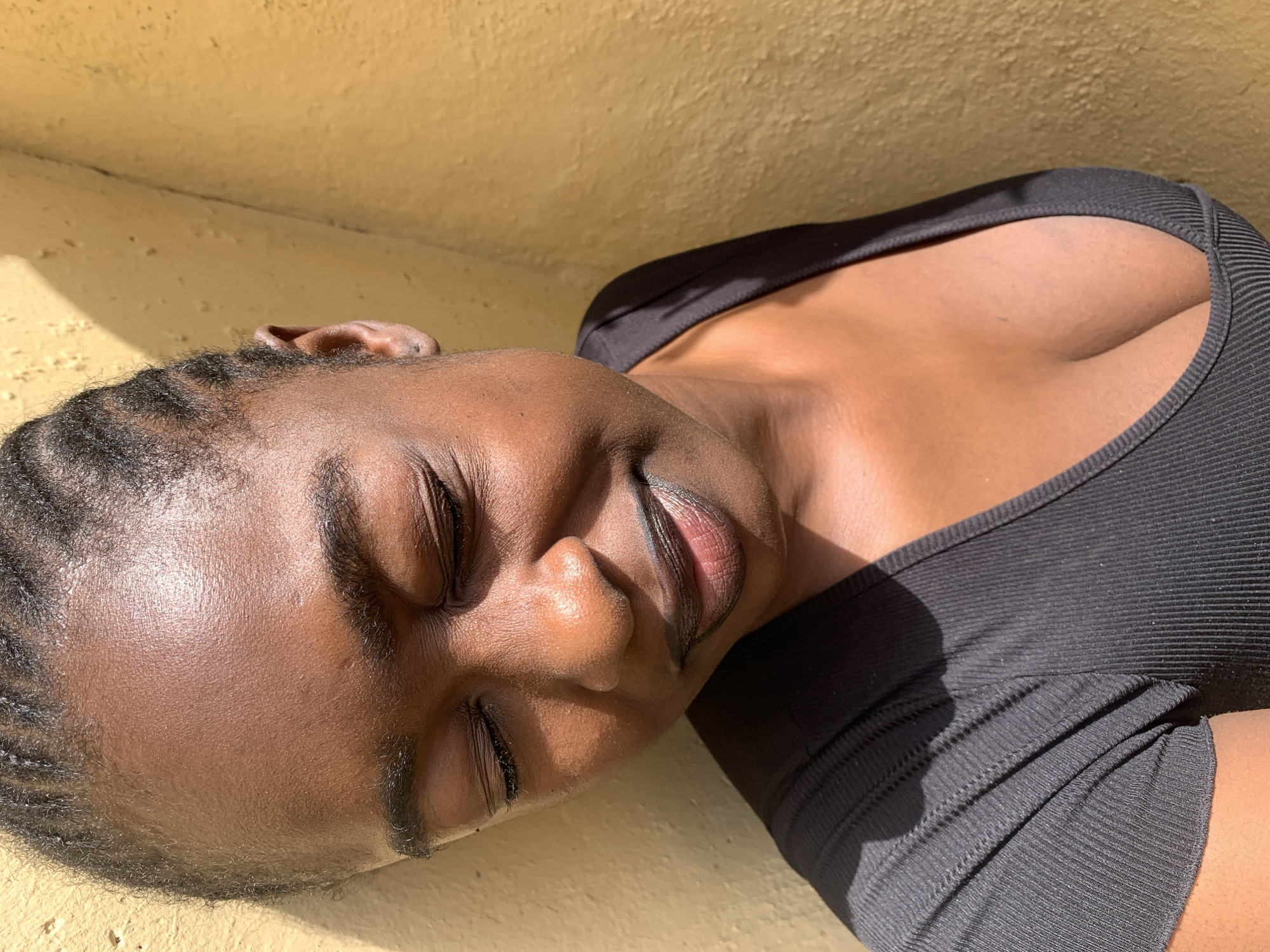 The image shows a close-up, horizontal shot of a young Black woman lying down, with her eyes closed and head tilted back. She is wearing a black, ribbed tank top. Sunlight illuminates her face and upper body, casting shadows and highlighting the texture of her skin. Her hair is styled in neat braids. The background is a textured, yellow wall, suggesting an outdoor setting. The lighting indicates it is daytime, possibly sunny. Her expression appears serene or peaceful. No text is visible in the image.