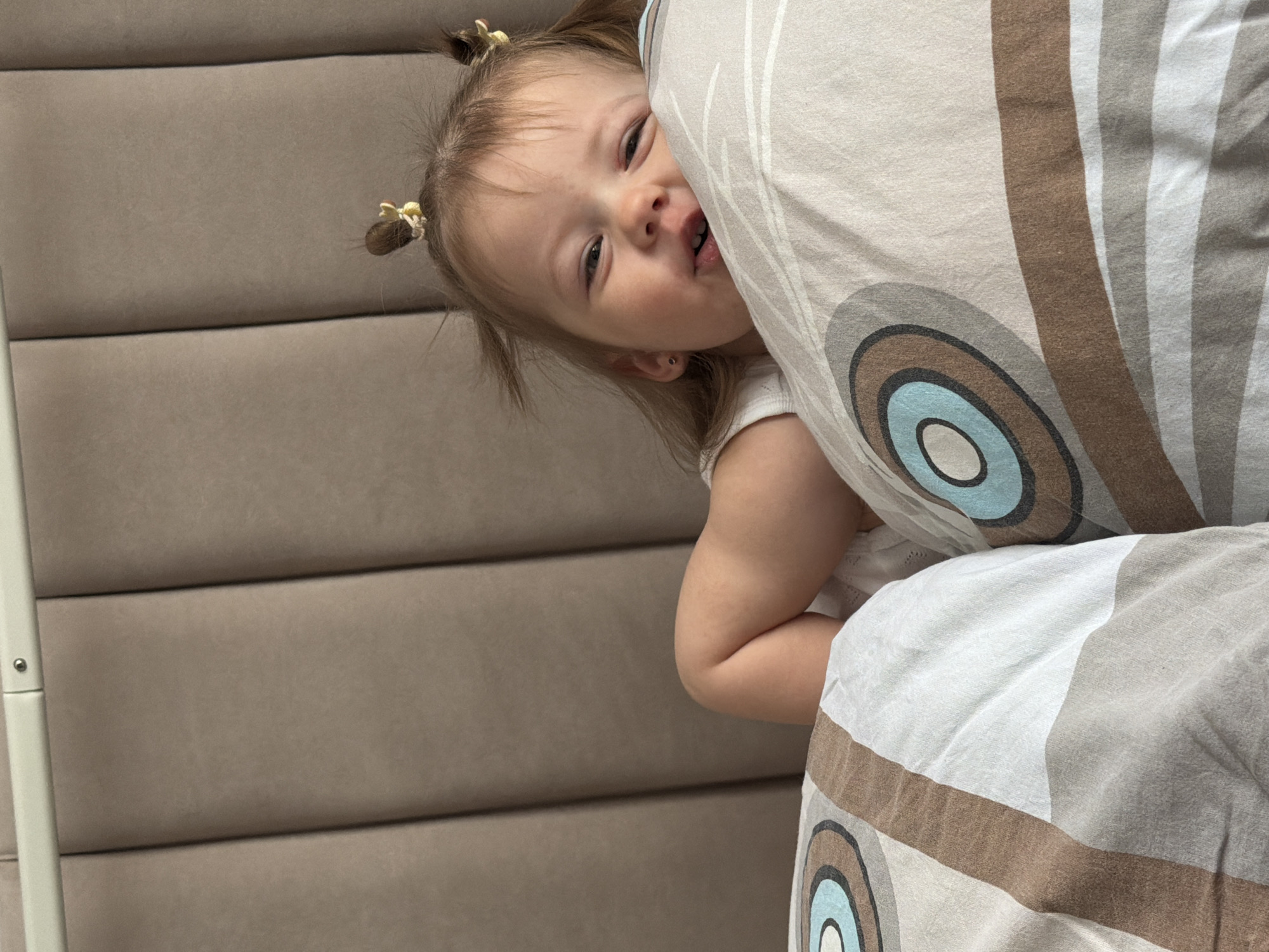A close-up, eye-level shot shows a young girl with blonde hair styled in two pigtails, partially obscured by a patterned pillow. The girl's face is visible, revealing a playful expression with an open mouth. The pillow features concentric circles in shades of blue, brown, and gray. The background features a tan, padded wall with vertical seams. The location and specific details regarding Reshetnikovo, Russia, are not discernible from the image.
