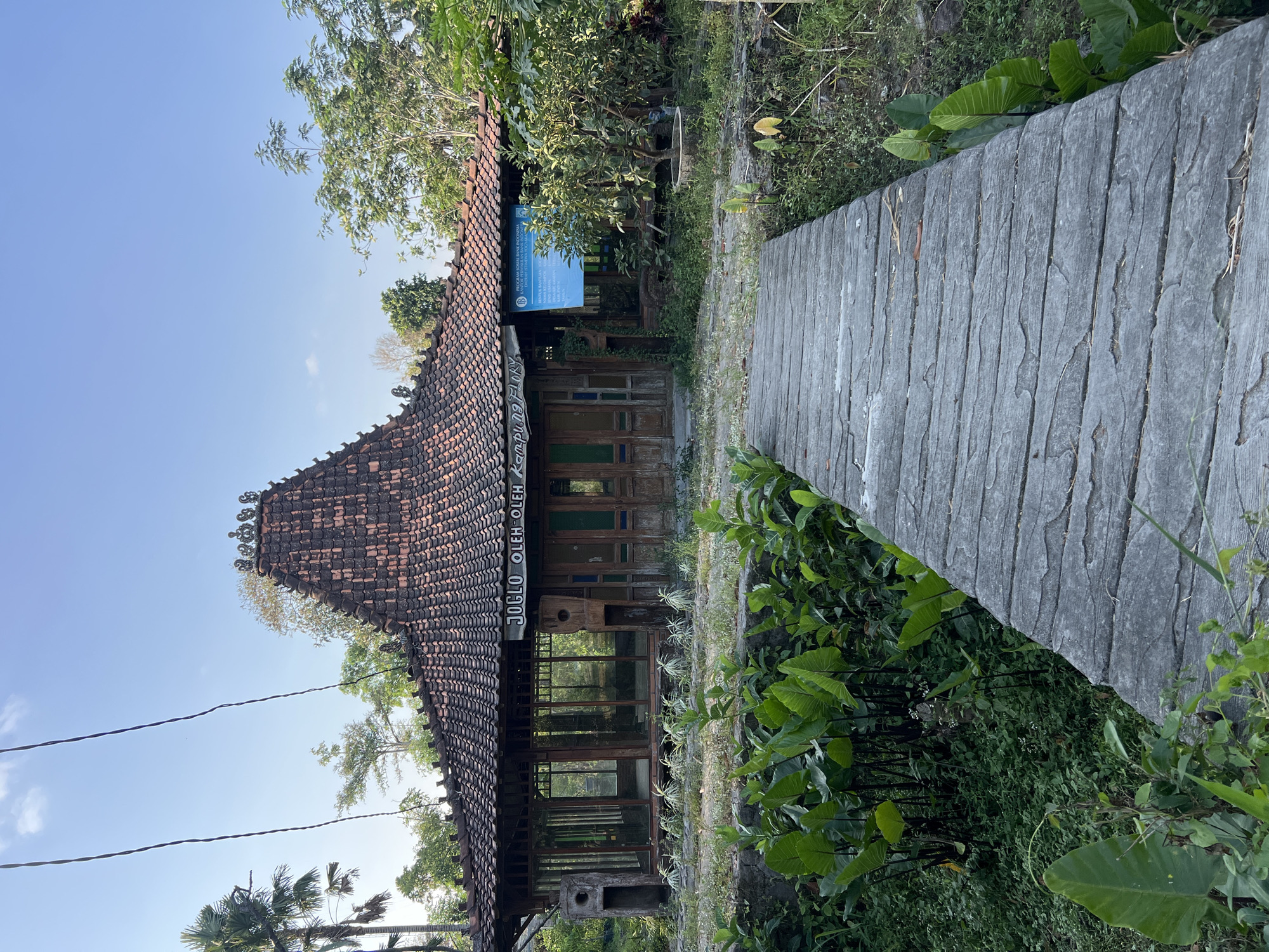 In the city of Tlogoadi, Indonesia, a traditional Javanese joglo building stands, adorned with intricately tiled roofing and decorative elements. A blue sign hanging from the building reads "PROGRAM SOSIAL BANK INDONESIA," indicating a social program sponsored by Bank Indonesia. The joglo, a heritage building, appears to be a shop selling souvenirs and local crafts, as suggested by the text "JOGLO OLEH OLEH KAMPUNG FLORY" on its facade. A concrete pathway leads to the entrance, flanked by lush greenery. The overall scene depicts a culturally rich and serene environment, with the joglo serving as a focal point amidst the natural beauty of the surroundings.