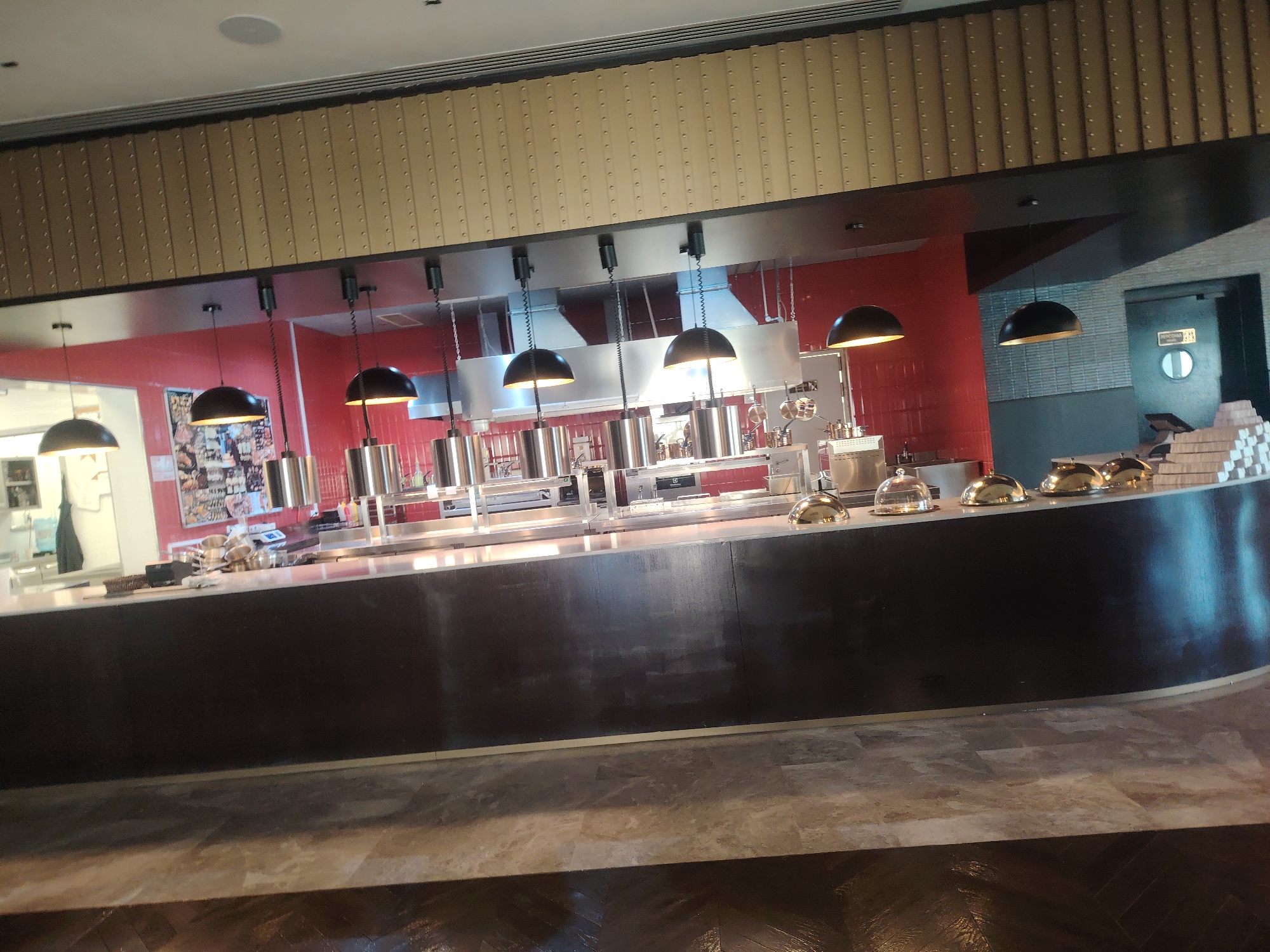 The image shows an interior view of a restaurant kitchen and serving area. In the foreground, there is a dark, curved counter with several reflective domes on top, possibly covering food. The background reveals a well-lit kitchen with stainless steel equipment, including stovetops, pots, and pans. Red tiled walls are visible behind the kitchen appliances. Several pendant lights hang from the ceiling above the counter and kitchen area. A doorway with a circular window and signage is present on the right side of the image. A stack of white boxes is also visible near the doorway. The flooring appears to be a combination of light-colored tiles and darker, reflective material. There are no people visible in the image. The scene suggests a commercial food service establishment, potentially a buffet or open kitchen concept. The city of Konigil, Uzbekistan is not depicted in the image.