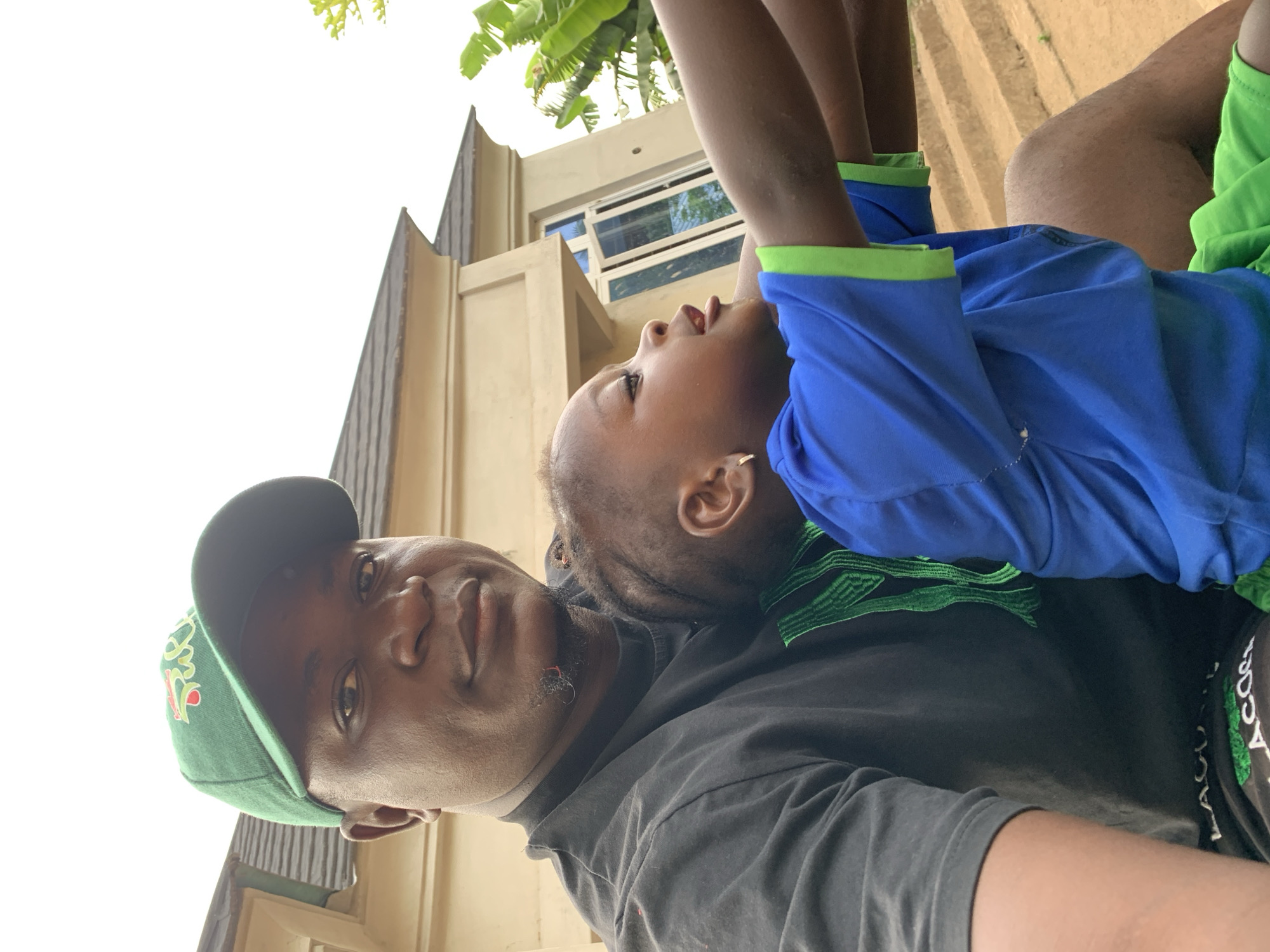 This outdoor portrait, rotated 90 degrees clockwise, captures a man and a young child engaged in a playful moment during the daytime in Akwanga, Nigeria.
In the foreground, an adult man is visible, wearing a dark grey or black t-shirt and a green baseball cap. He has a short goatee and looks directly at the camera with a neutral, calm expression. He is holding a young child, who appears to be playfully suspended upside down, with their head near the man's left shoulder. The child is looking upwards and slightly to their right, away from the camera, displaying a calm and curious expression. They are dressed in a bright blue t-shirt with lime green trim on the collar and sleeve cuffs, and a small earring is visible in their left ear. The child's arms are bent upwards, adding to the playful posture.
The background reveals a bright, uniformly white sky, suggesting an overcast day, which dominates the upper left portion of the image. Behind the subjects, a light-colored building with visible windows indicates a residential or communal setting. Green foliage, possibly banana leaves, can be seen in the upper right, characteristic of a tropical environment.
Notable details include the text on the man's green cap, which is stylized and not fully legible. More distinctively, on the man's dark shorts or pants, white text reads "LAGOS" and "CRO" (possibly part of "CROC"), set against a green pattern. The coordinated green accents in both the man's attire (cap, shorts) and the child's outfit (shirt trim) create a subtle visual connection between them. The image conveys a sense of warmth and lighthearted interaction.