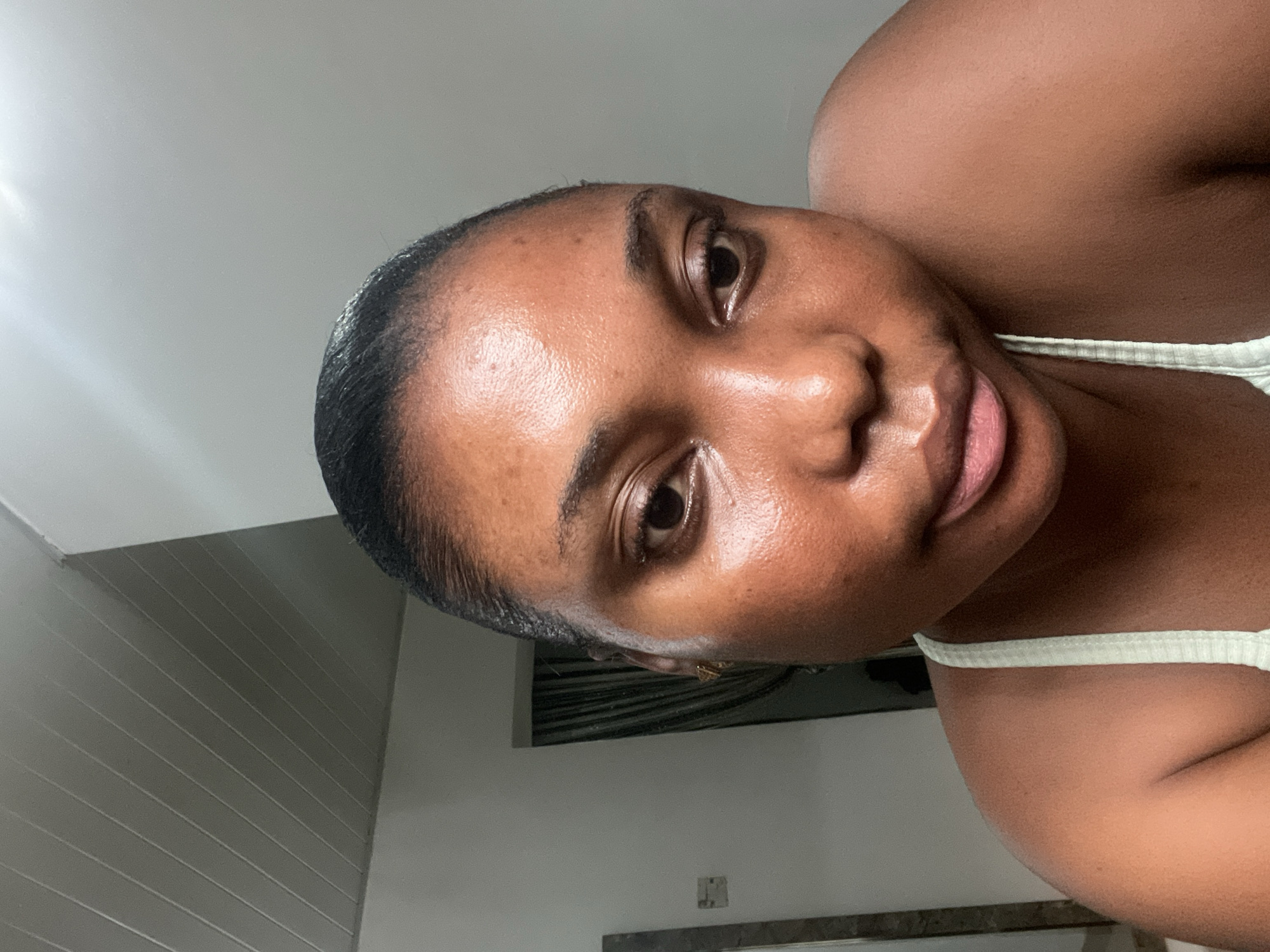 A close-up photograph shows a woman with dark skin facing the camera. She has dark hair slicked back from her forehead. She is wearing a light-colored tank top. The background includes a white ceiling and part of a wall with vertical paneling. Behind her, there is also a dark rectangular object, possibly a piece of artwork or a mirror. The setting appears to be indoors. There is no specific evidence to determine if the image was taken in Abuja, Nigeria.