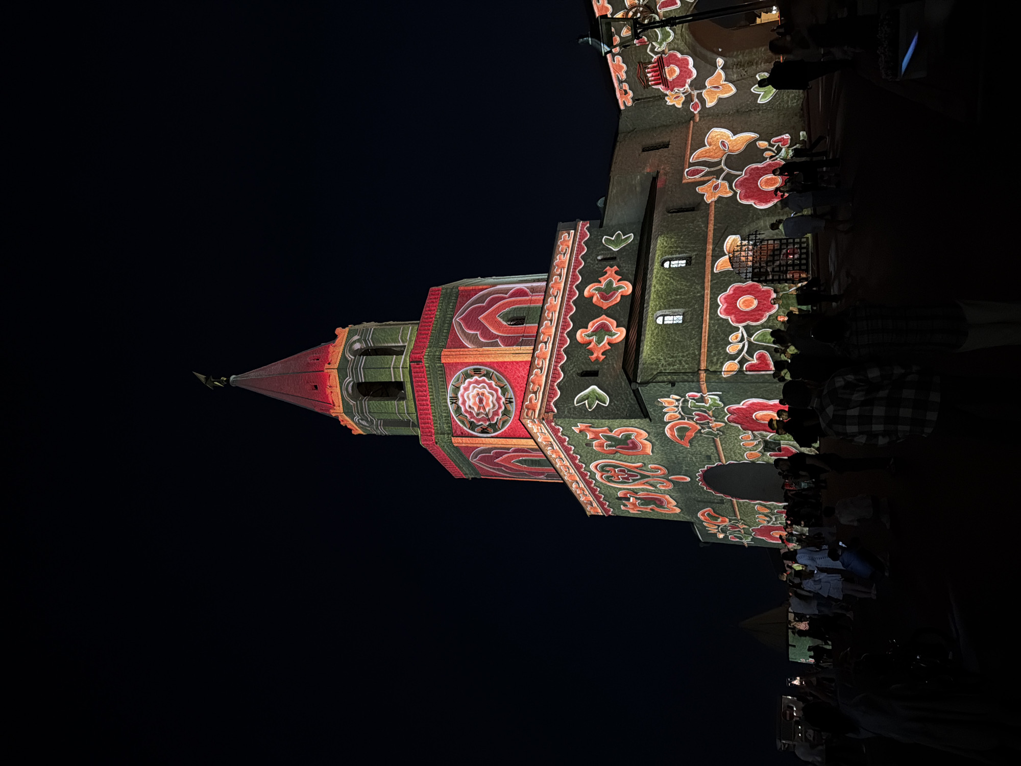 At night, a tall, ornate tower is illuminated with vibrant, colorful projections. The tower, likely part of the Kazan Kremlin, is adorned with patterns resembling flowers, leaves, and geometric designs. A clock face is visible on one side of the tower. Silhouetted figures of people stand near the base of the tower, seemingly observing the light display. The background is dark.