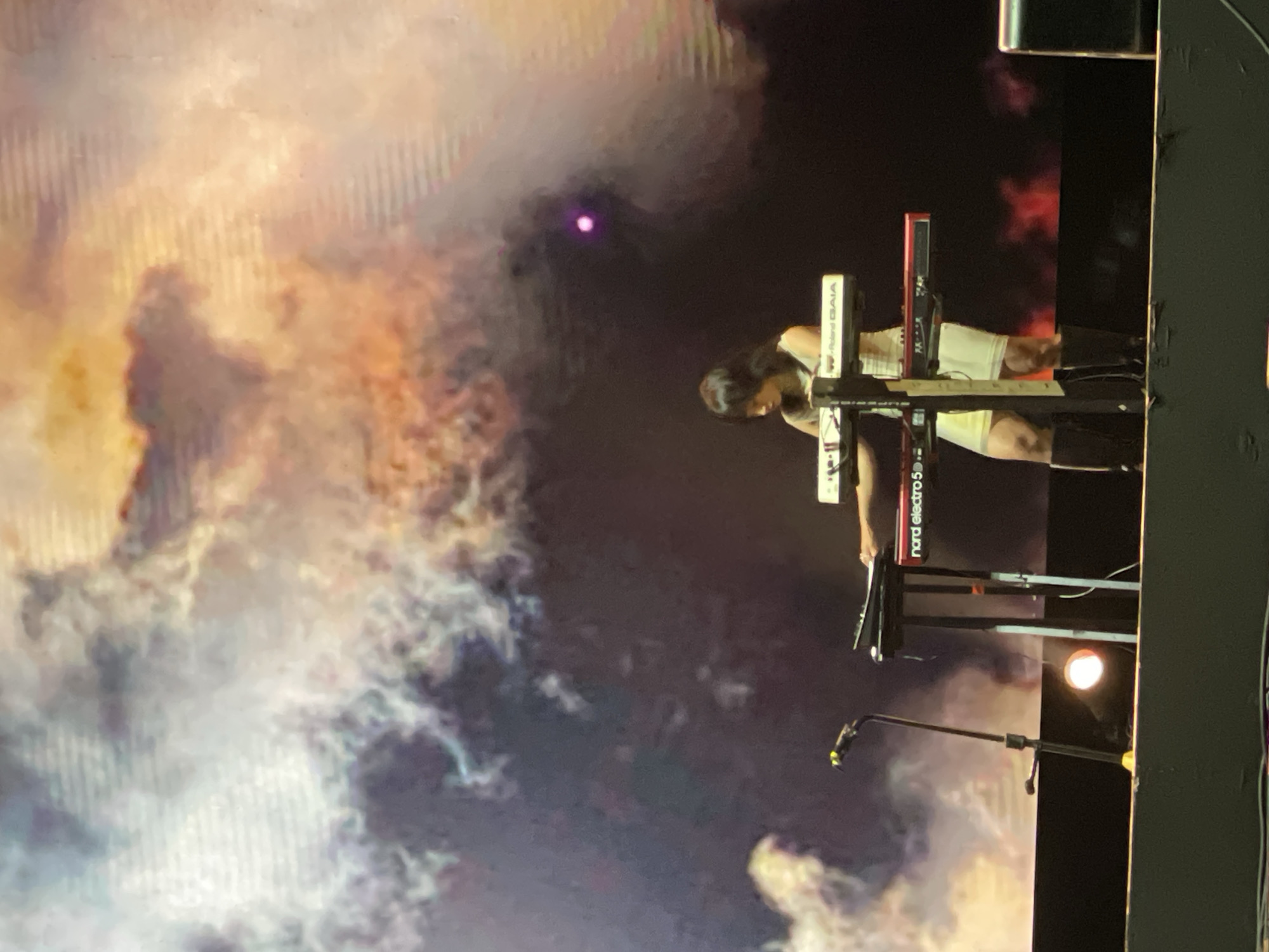 The image shows a performer playing keyboards on a stage. The performer, with dark hair, is wearing a white top and shorts. The keyboards include a Roland GAIA and a Nord Electro 5. A microphone stand is positioned near the keyboards. The backdrop features a cloud-like or sky-like pattern. The image was likely taken during a performance in Salembaran, Indonesia, indicated by the source mentioning this location.