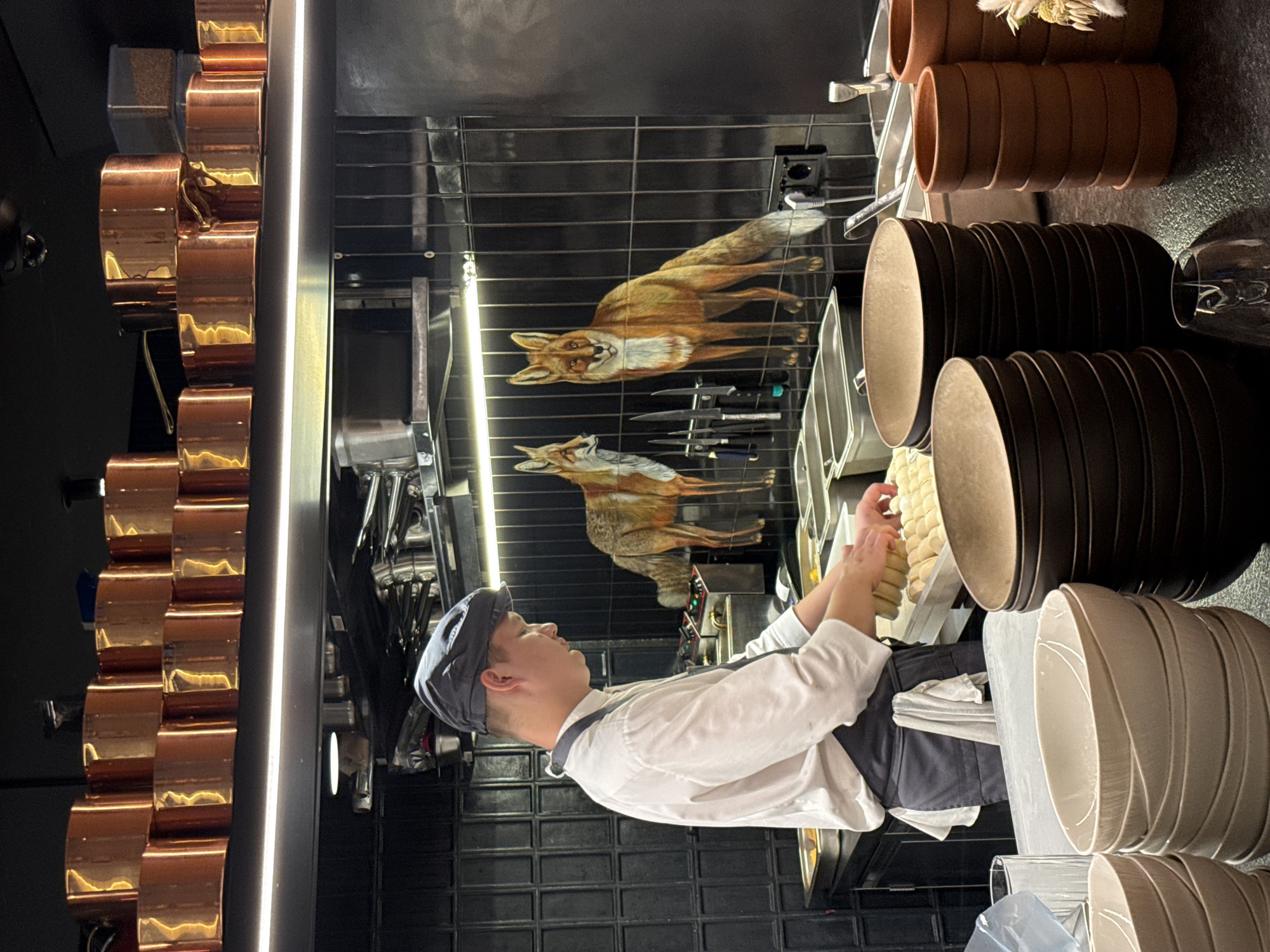 The image depicts a chef in a kitchen, wearing a white chef's coat and a dark cap. He is working with small, round food items, possibly dough or bread rolls, which are arranged in rows on a metal tray. The kitchen appears to be well-equipped, with copper pots hanging on the left side of the frame and stacks of dark plates on the right.
A notable detail is a reflection on the black tiled wall behind the chef. This reflection shows two paintings of foxes, one larger and more detailed than the other, positioned as if they are within the kitchen space. There are also knives mounted on a magnetic strip visible in the reflection.
The lighting suggests an indoor setting, likely during operational hours given the presence of the chef and the reflective surfaces. The scene is orderly and professional, hinting at a restaurant or high-end culinary establishment. No text is visible in the image. The overall impression is one of focused work and artistic elements incorporated into the kitchen environment. The location context is Moscow, Russia.