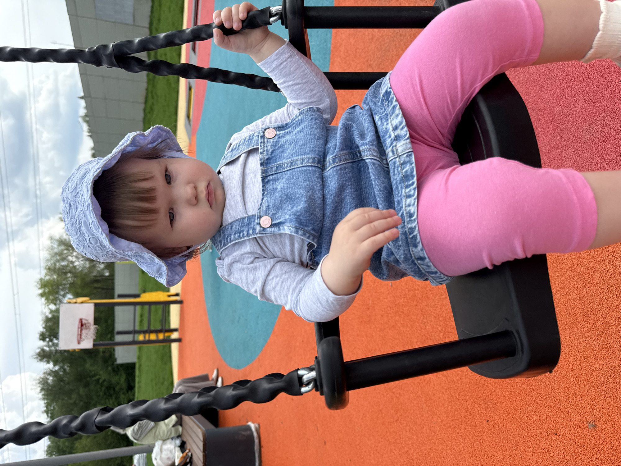A young child wearing a light blue sun hat, a light gray long-sleeved shirt, a denim dress, and pink leggings is sitting on a black swing at a playground. The playground has a multicolored surface with sections of red, blue, and orange. In the background, there is a basketball hoop and trees. The location is Moscow, Russia.