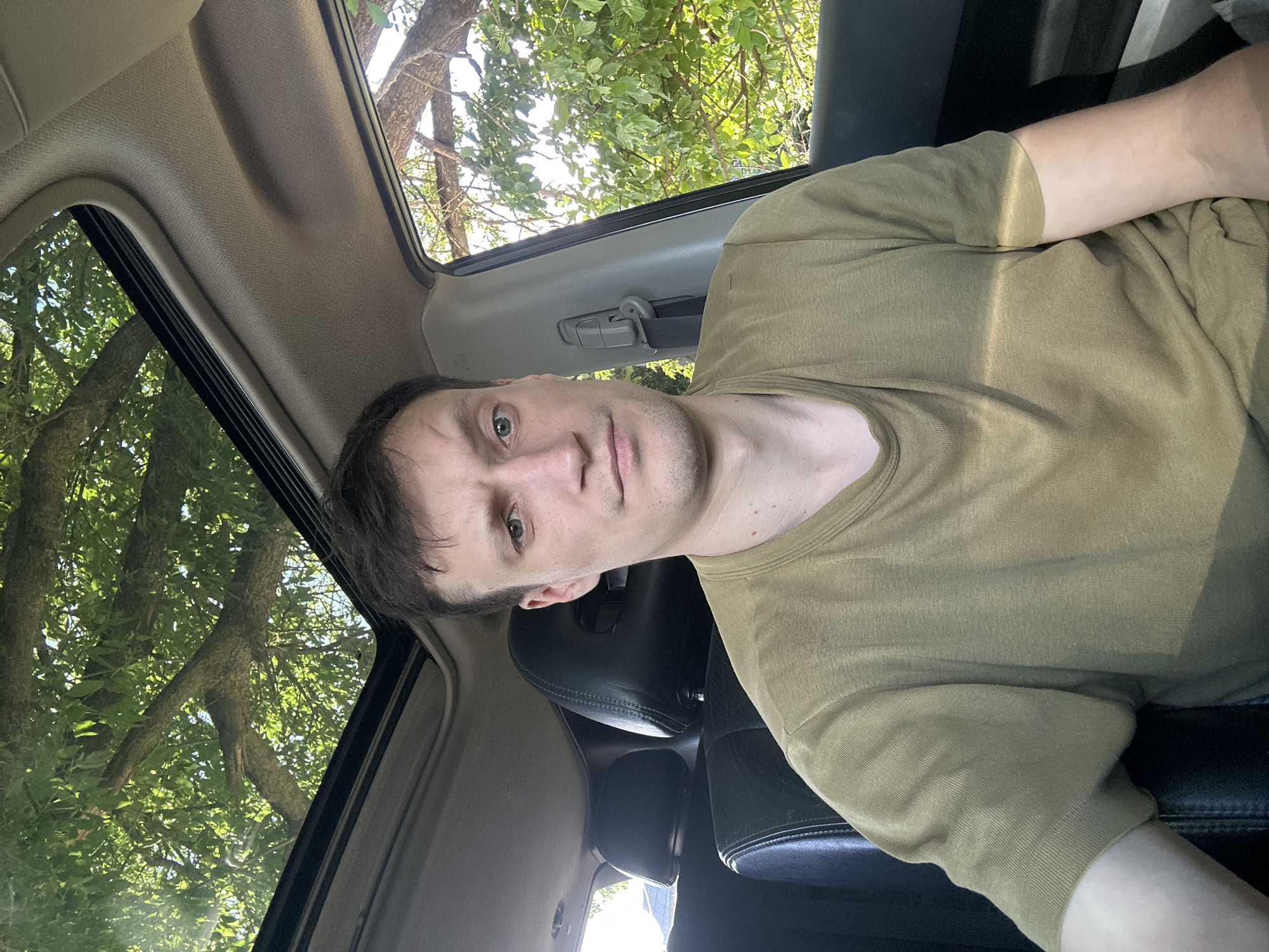 A man is sitting in the driver's seat of a car. He has short brown hair, light skin, and is wearing a plain olive green t-shirt. The car has black leather seats and a sunroof. Through the sunroof and the open window, trees and foliage are visible, indicating an outdoor location with greenery. The background suggests a sunny day. The photo appears to be a selfie taken inside the car. The location in Makiv, Ukraine, is not determinable from the image alone.
