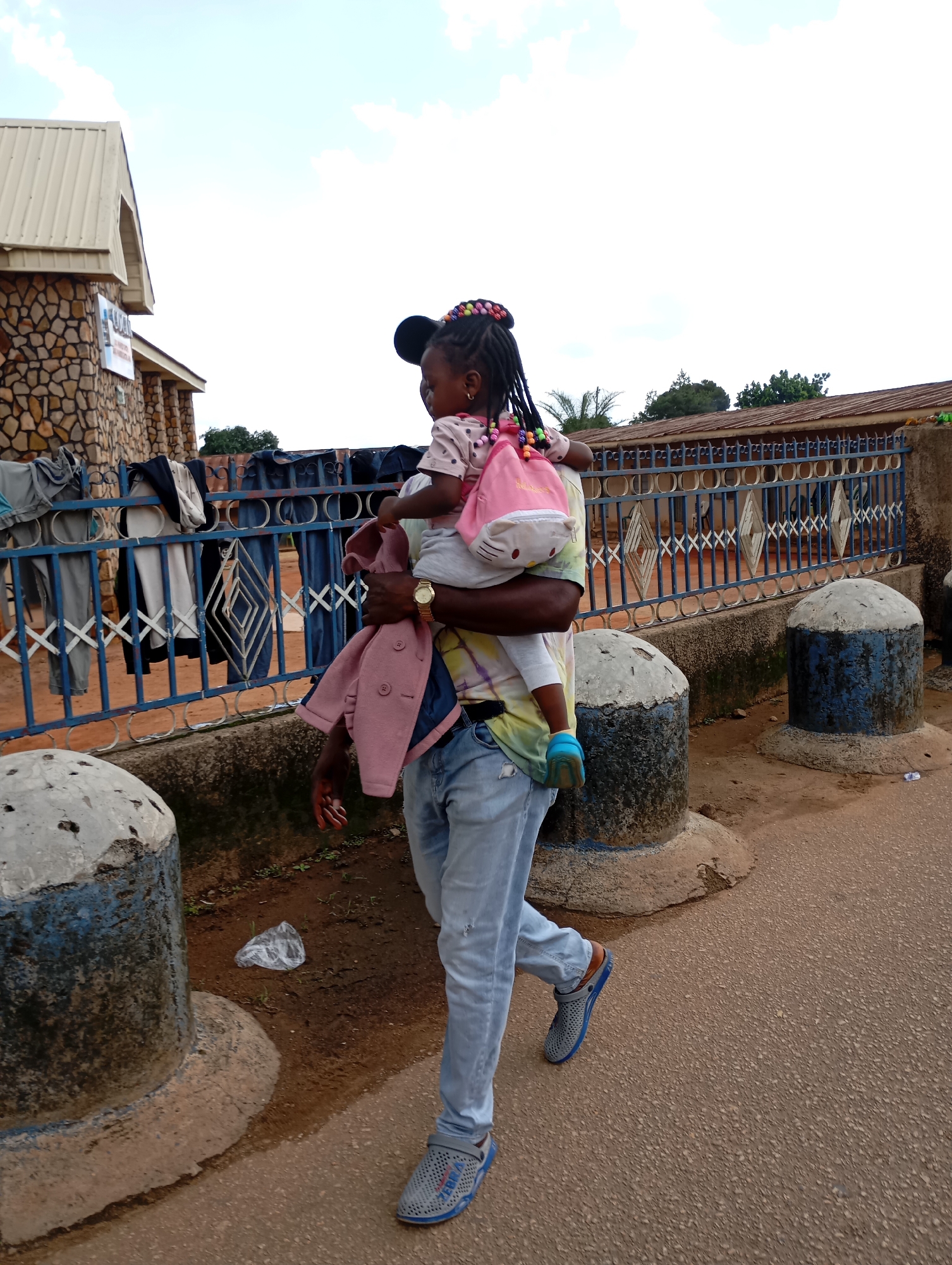 A man carries a young child while walking on a dirt road in Jos, Nigeria. The man is wearing a tie-dye shirt and blue jeans, and he has a watch on his left wrist. The child is wearing a pink jacket and a pink backpack. In the background, there is a building made of stone and a blue metal fence with clothes hanging on it. There are also two concrete pillars on the side of the road. The sky is cloudy.