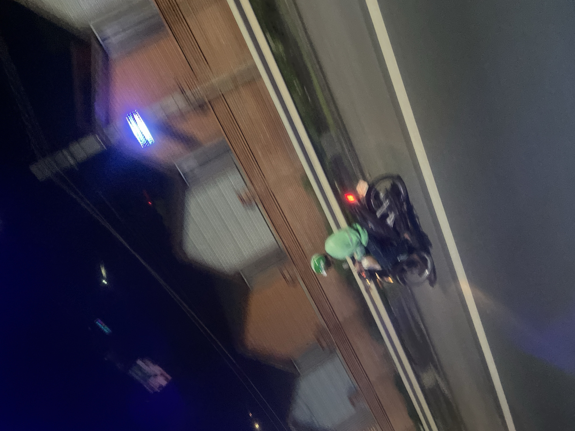 This image captures a night scene in Binh Thuan Ward, Vietnam, with motion blur indicating movement. A person wearing a green helmet and a bright green jacket is riding a motorcycle on a road. The motorcycle has a visible red taillight. In the background, a brick wall borders buildings with white roofs. Above the buildings, a bright blue LED light fixture is illuminated. The scene appears to be an urban or suburban setting at night.