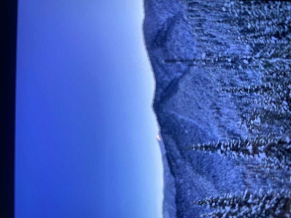 A winter landscape fills the right portion, showing snow-covered mountains and trees. To the left, a bright blue sky suggests a clear day. The contrast between the vibrant sky and the snow-laden terrain creates a striking visual divide. The overall orientation of the image is rotated 90 degrees clockwise.