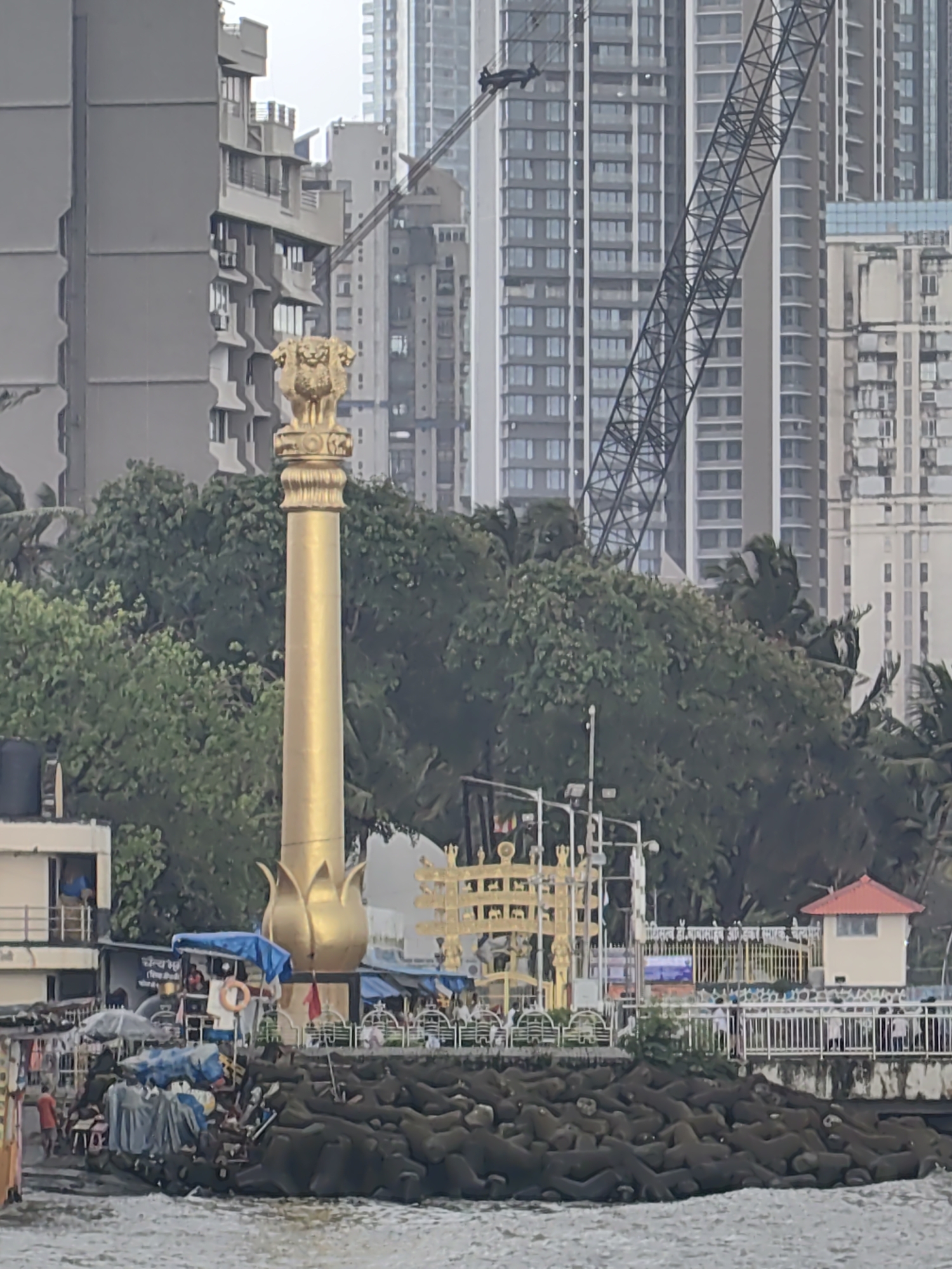 An exterior, eye-level shot captures a golden Ashoka Pillar monument featuring four lions at its apex, positioned on a rocky shore in Mumbai, India. The monument stands tall against a backdrop of lush green trees and a cluster of tall, modern buildings under an overcast sky. In the foreground, the sea laps against a breakwater constructed of large, dark, angular concrete blocks. Various structures and facilities are visible on the shore, including a building with a red-tiled roof, a white fence, and what appears to be market stalls or kiosks with blue tarpaulins. A yellow, ornate gateway stands near the base of the monument. Several people are present on the shore, some near the stalls and others by the fence, suggesting a public area or tourist attraction. The presence of construction cranes in the background indicates ongoing development in the urban landscape.