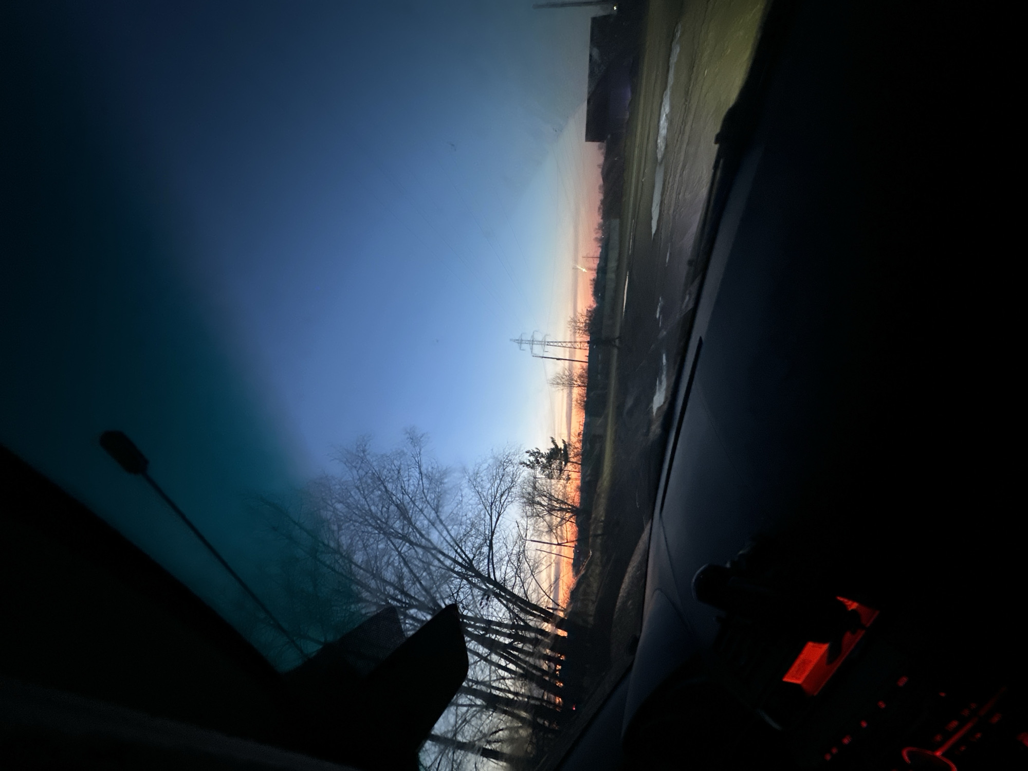 The image depicts a sunset observed from the interior of a car. The sky transitions from blue at the top of the frame to orange near the horizon. Silhouetted trees and power lines are visible against the bright sky. Reflections of streetlights can be seen on the windshield. A building and a wet parking area can be seen through the windshield. The dashboard and interior components of the car are visible, though largely obscured by shadow. The location is inferred to be near Chaikovsky, Russia, based on the prompt. No people are visible.