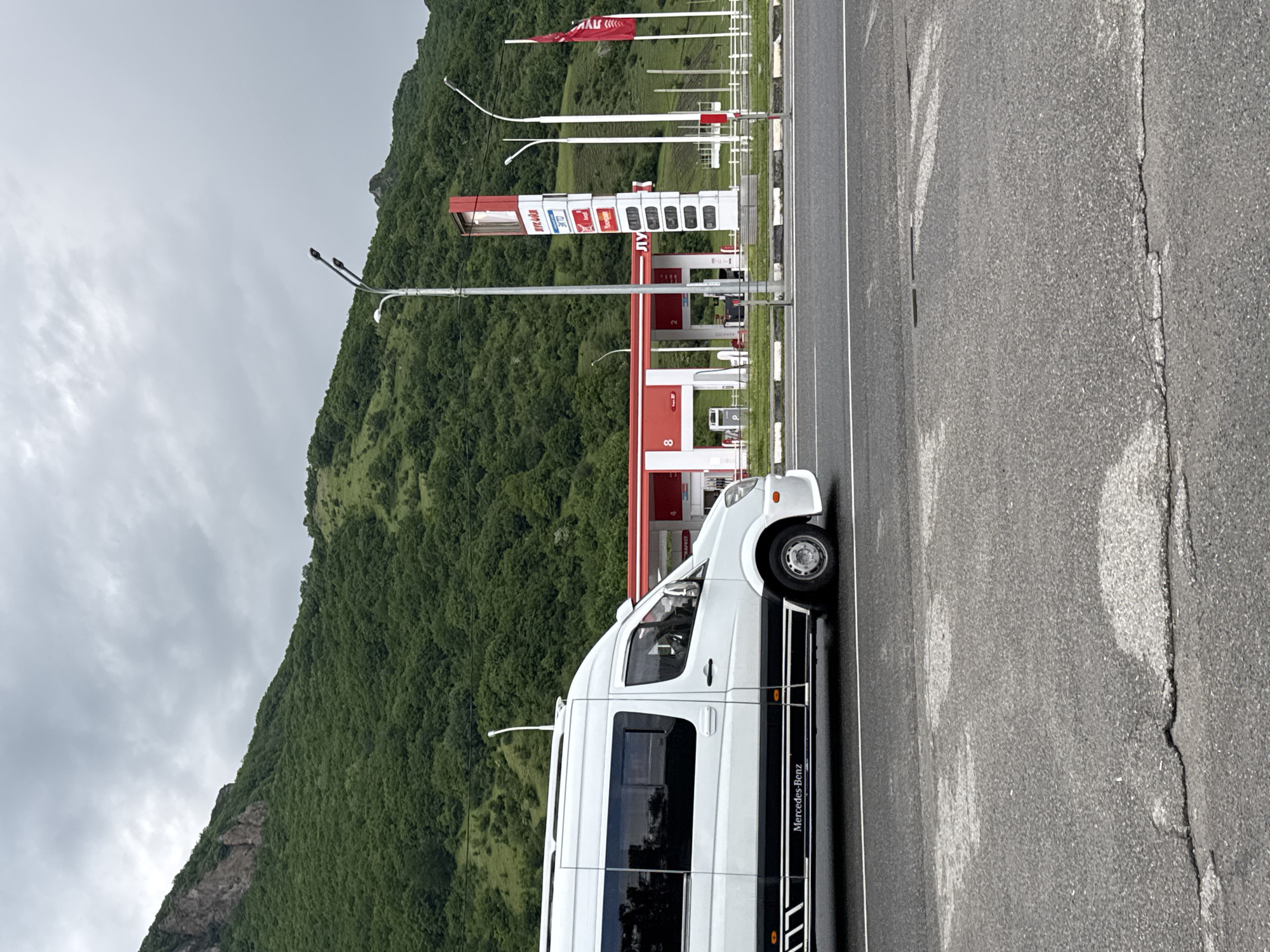 A white Mercedes-Benz Sprinter van is parked on the side of a road with a gas station in the background. The gas station has a red and white color scheme and a tall price display. Mountains covered in green trees rise behind the gas station under a cloudy sky. The road in the foreground is asphalt with white lane markings and some cracks. The scene appears to be in a mountainous region, possibly near Karachayevsk, Russia, given the architecture and landscape.