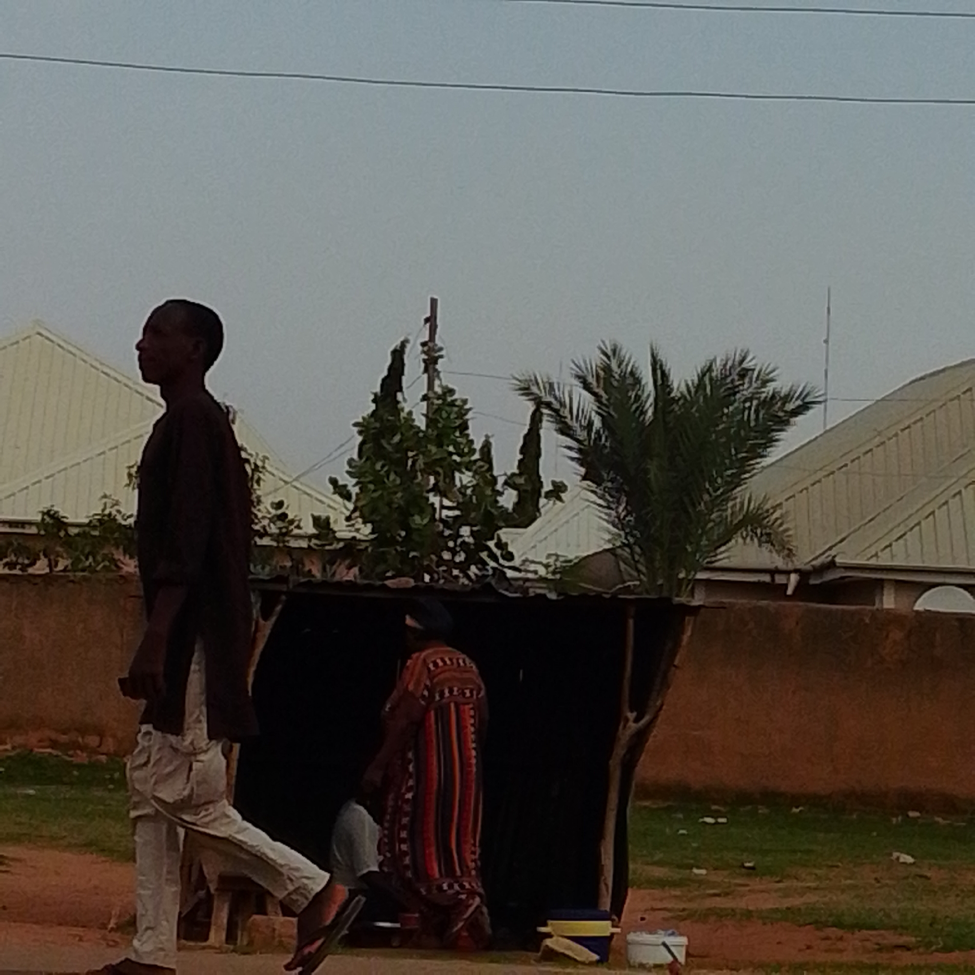 The image depicts a scene in Gombe, Nigeria, with two individuals visible. In the foreground, a man is walking from left to right, wearing a dark long-sleeved shirt and light-colored trousers. His profile is visible as he walks past a makeshift stall. Behind him, within the stall, a person is seated or standing, wearing a colorful, long patterned garment, possibly a dress. The stall appears to be constructed with wood and a dark covering material. In the background, there are buildings with light-colored, sloped roofs and lush green vegetation, including palm trees and other leafy plants. A power line is visible stretching across the sky, and the overall lighting suggests it is either early morning or late afternoon. The ground is a mix of dirt and patchy green grass.