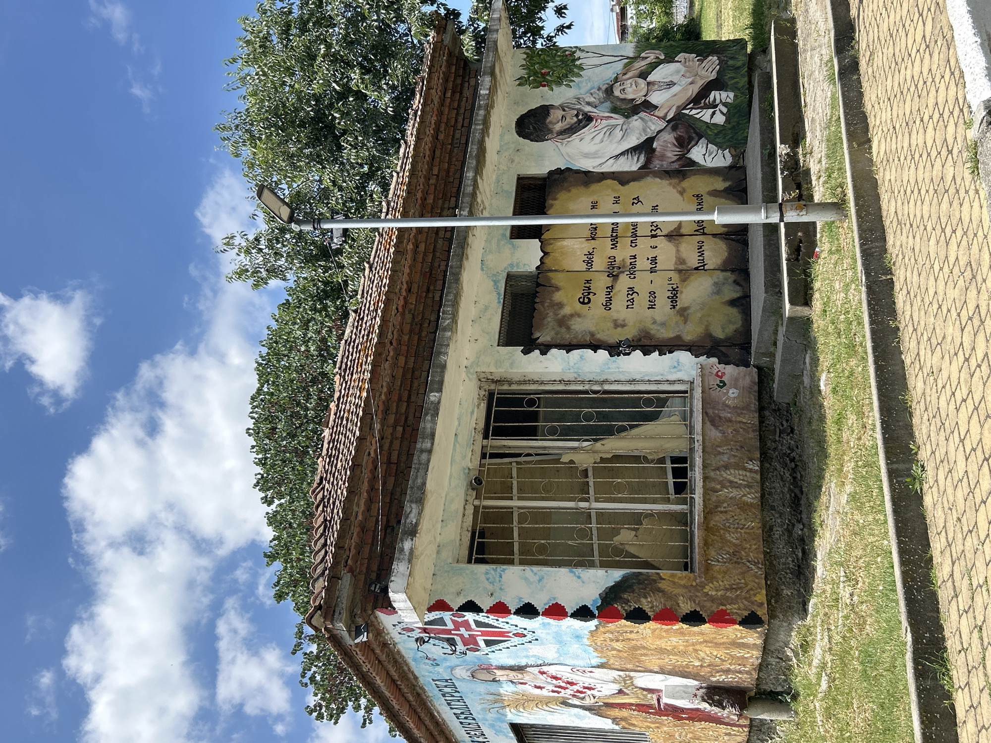 The image shows a mural painted on the exterior of a small, weathered building in Aheloy, Bulgaria. The mural depicts traditional Bulgarian cultural imagery. On the left side, a woman in traditional attire holds a sheaf of wheat, with Bulgarian script above her. A stylized flag design is also present. The central part of the mural features a quote in Bulgarian text written on a parchment-like background, alongside a depiction of a man and a child. The man, with a beard and wearing traditional clothing, is shown placing his hand on the child's head. A small tree with red fruit grows next to them. The building itself has a tiled roof, barred windows, and is situated on a paved area with grass and a sidewalk. A streetlamp stands in front of the mural. The sky above is blue with scattered clouds.
