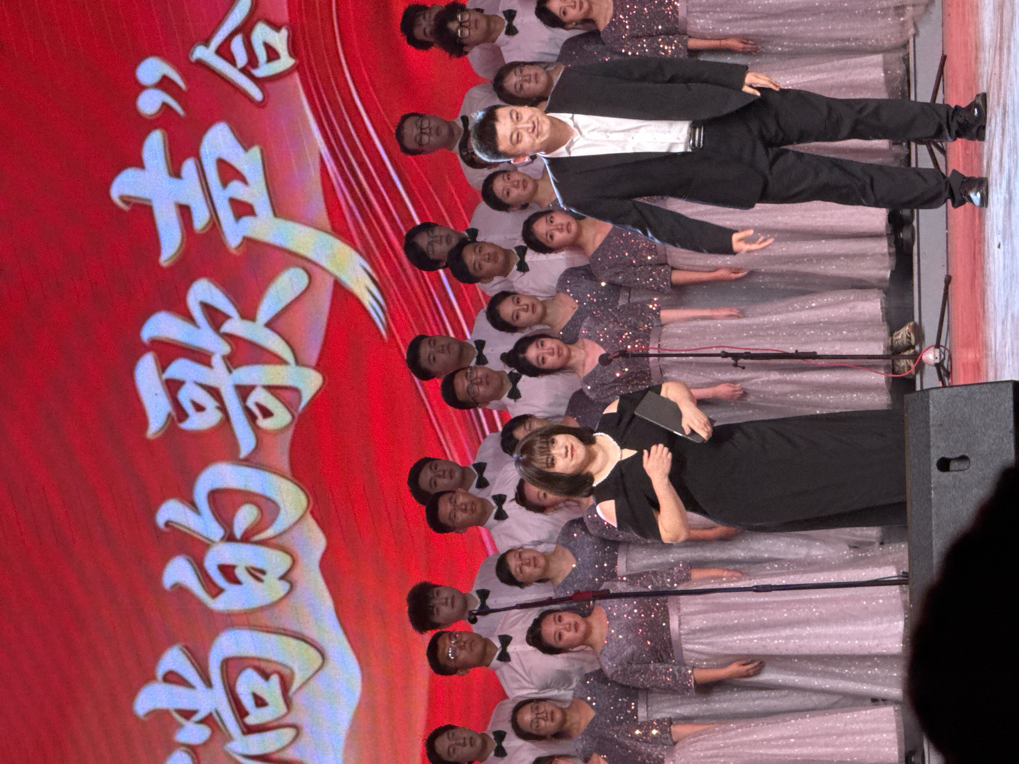 This vertical image captures a large choir performing on an indoor stage, likely during a concert or musical event in Guta, China. The ensemble is composed of both men and women arranged in several rows.
In the foreground, a woman in a black dress holds a black book or folder, standing next to a microphone, seemingly conducting or acting as a soloist. Behind her and to the side, a man in a black suit, white shirt, and black tie stands prominently, possibly as a conductor or lead vocalist, looking upwards with a faint smile.
The choir members are formally dressed: the men wear white button-up shirts with black bow ties, while the women are in shimmering light purple or pink dresses. They appear focused and attentive during the performance. Several microphones on stands are visible among the performers.
The backdrop is a vibrant red screen adorned with large, stylized white and light-colored Chinese characters arranged vertically. The visible characters, reading from top to bottom, are "会", "音", "乐", "的", "青", and "春". This translates to "会音乐的青春" (Huì Yīnyuè de Qīngchūn), meaning "The Youth of Music Gathering" or "Youth Who Can Play Music," suggesting a youth music or choir concert.
The lighting is consistent with an indoor stage environment, highlighting the performers and the backdrop. The overall atmosphere conveys a sense of formal presentation and musical artistry.