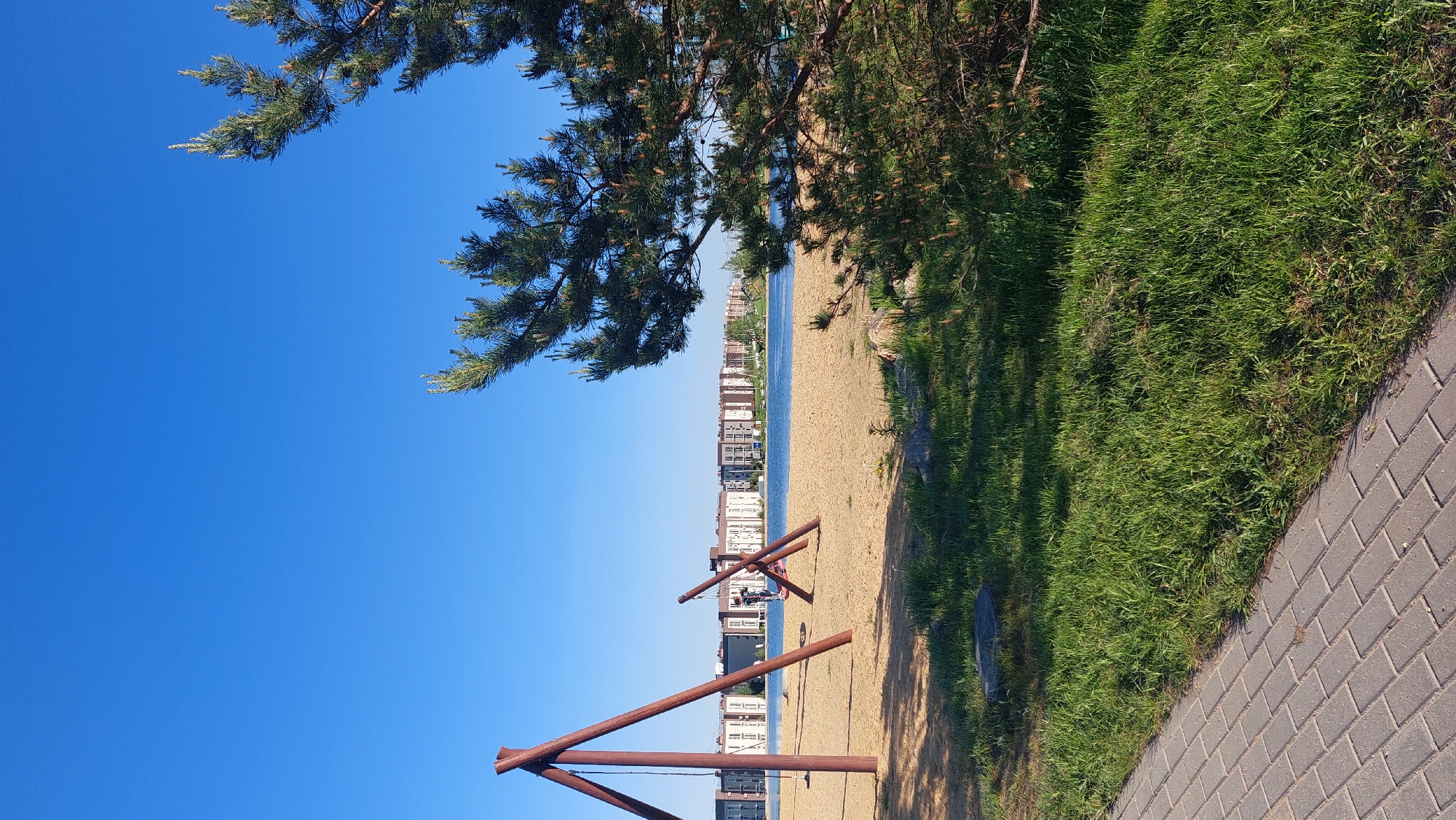 The image presents a view of a beach, likely situated on the shore of a body of water, with a cityscape visible in the background. Based on the cityscape and the presence of the wooden playground structure, the location is likely the beach on Smolino Lake in Chelyabinsk, Russia.
The foreground contains a wooden playground structure made of logs, with sand underneath and around it. Adjacent to the sandy area is a section of green grass, leading to a paved walkway made of interlocking pavers. Overhanging branches and foliage are visible at the top of the frame, partially obscuring the sky. The sky is clear and blue.
In the background, a line of buildings suggests an urban setting, lining the opposite shore of the body of water. The water itself appears calm and reflects the sky.