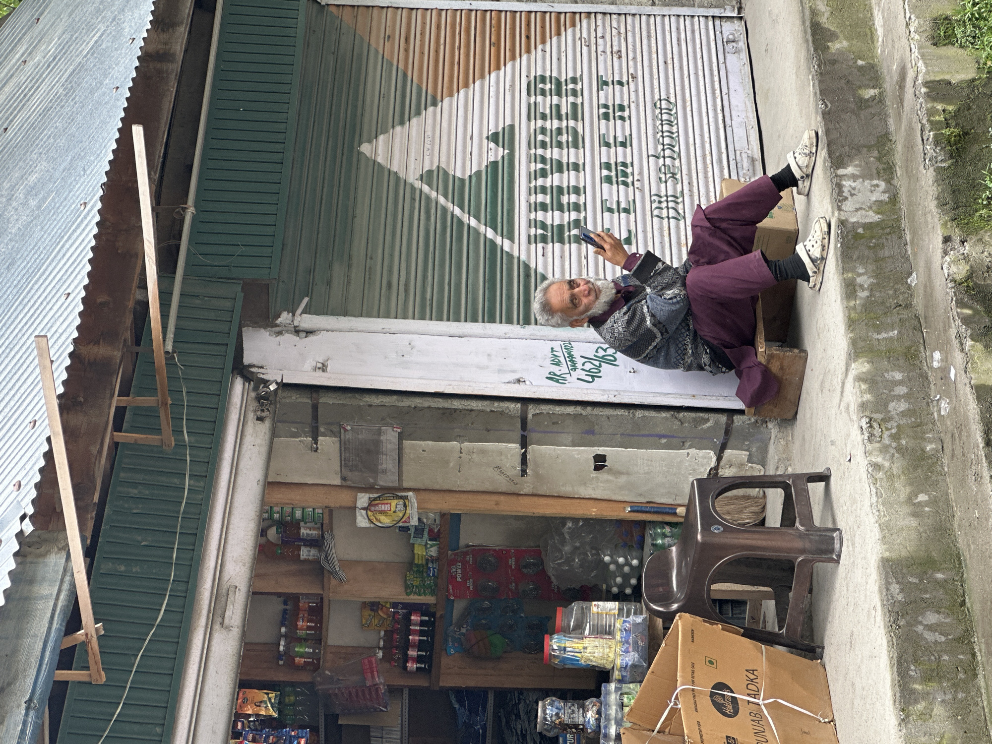 An elderly man with a white beard and grey hair is sitting on a small wooden bench outside a shop in Kullan, India. He is holding a smartphone in his right hand and appears to be looking at it. He is wearing a dark patterned sweater over a maroon shirt and maroon pants, with black socks and white Crocs. The shop has a green corrugated metal facade with a roller shutter that is partially rolled down. A large white and green triangular logo with the word "KHYBER CEMENT" is painted on the shutter, along with the Hindi phrase "Dil se bannao". To the left of the man, there is a brown plastic stool, a cardboard box labeled "PUNJAB TADKA", and shelves stocked with various goods, including bottles of drinks and packaged items. The shop appears to be a small convenience store. The ground in front of the shop is concrete, with some moss and dirt. The overall environment suggests a roadside stall in a town or village.