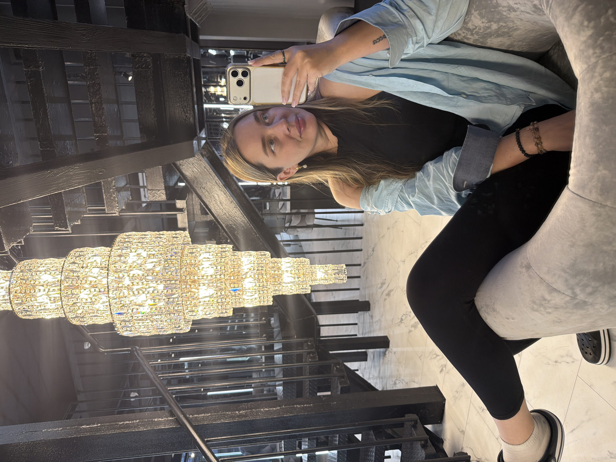 A woman sits in a gray upholstered chair. She wears black leggings, a black top, and a light blue shirt. She holds a white smartphone to take a photo. Behind her is a black metal staircase with a large chandelier hanging above it. The location is Moscow, Russia. The floor has a light-colored marble pattern.