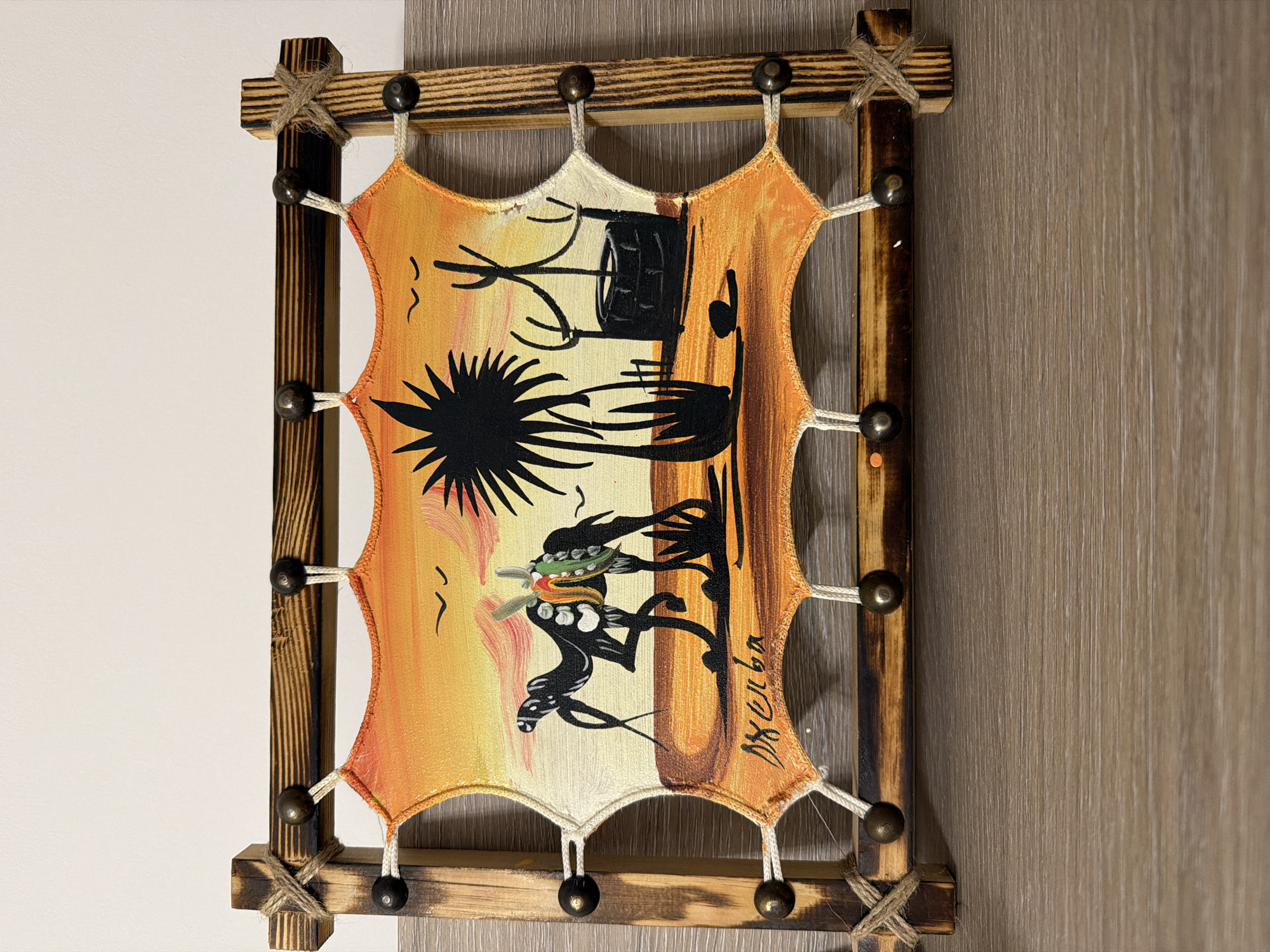 The image shows a framed, hand-painted piece of art hanging against a wall. The frame appears to be made of wood with a darker, possibly stained or burned finish. Twine is wrapped around the corners where the wood pieces meet. The artwork itself is stretched taut within the frame and features a scene depicting an African-inspired landscape. There's a silhouette of a well, a palm tree, and stylized figures of people, one of whom appears to be carrying something on their head. The colors in the painting are predominantly warm tones, suggesting a sunset or desert environment. The signature "Джела" is visible in the lower right corner of the artwork.