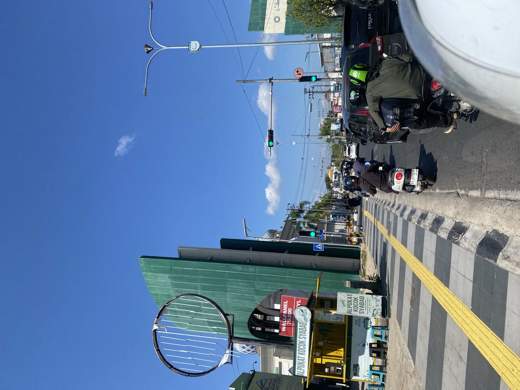 The image depicts a bustling street scene in Jawa, Indonesia, under a clear blue sky. In the foreground, a person is riding on the back of a motorcycle, looking forward at the traffic ahead. They are wearing a green shirt and a helmet with the "Gojek" logo. The motorcycle is in motion, indicating movement through the city.
The street is lined with various establishments, including a bright yellow food stall advertising "ALPUKAT KOCON SYABAB" (Avocado Shake Syabab). Behind the food stall, a large, green, corrugated metal structure stands tall, with a circular, wire-frame sculpture attached to its front. Further down the street, buildings with domed roofs, characteristic of mosques, are visible. Traffic signals, some displaying green lights, indicate the flow of vehicles. Cars and numerous motorcycles are present on the road, suggesting a busy urban environment. The sidewalk features a distinctive yellow tactile paving strip, designed for visually impaired pedestrians. The overall atmosphere is one of daily urban life, with people commuting and engaging in commerce.