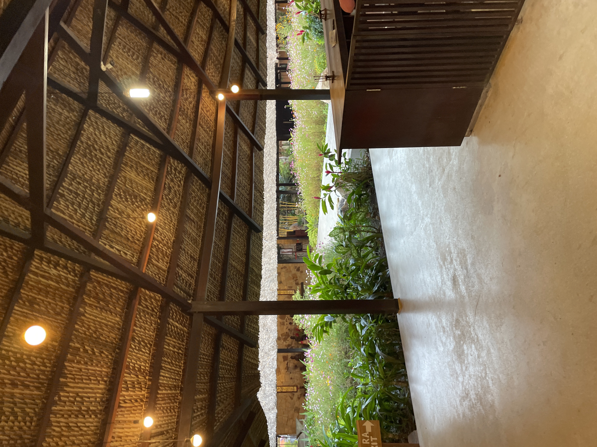 The image depicts a partially obscured view of a building exterior. The left side of the frame is dominated by a steeply pitched thatched roof, supported by dark wooden beams. Small, spherical lights are attached to the underside of the roof.
To the right of the roof, a strip of greenery is visible, featuring a variety of plants and flowers. Beyond this, a wall constructed from light-colored stone is seen. Further to the right is a dark brown, cabinet-like structure, set against a light-colored concrete floor.
The location appears to be Nha Trang, Vietnam, based on the architectural style and the presence of tropical foliage.