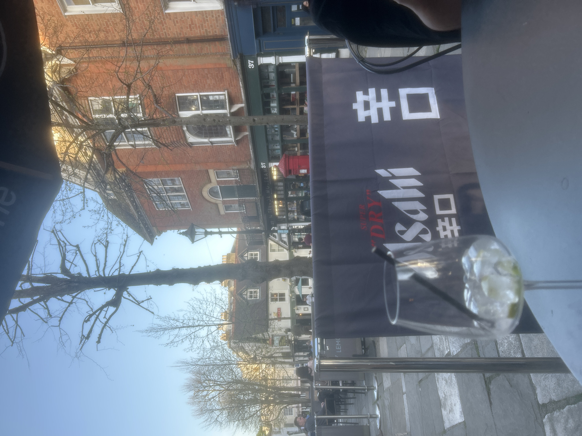 The scene shows an outdoor seating area, potentially a restaurant or bar. In the foreground, a drink in a glass with a straw is visible. A banner with Japanese characters and the word "Asahi" is also prominent. Looking beyond the seating area, the buildings on either side of the street are visible. One building is made of red brick, with multiple windows. The building on the other side of the street has a blue storefront, with the number 37. On the left, there is a building and its address appears to be "37 M TRE OR MOTRA M37". There are trees lining the street, many of which are bare of leaves. People can be seen sitting and walking along the street. The sky is light blue, suggesting it is daytime. A sign for "The Duke" is visible in the background.