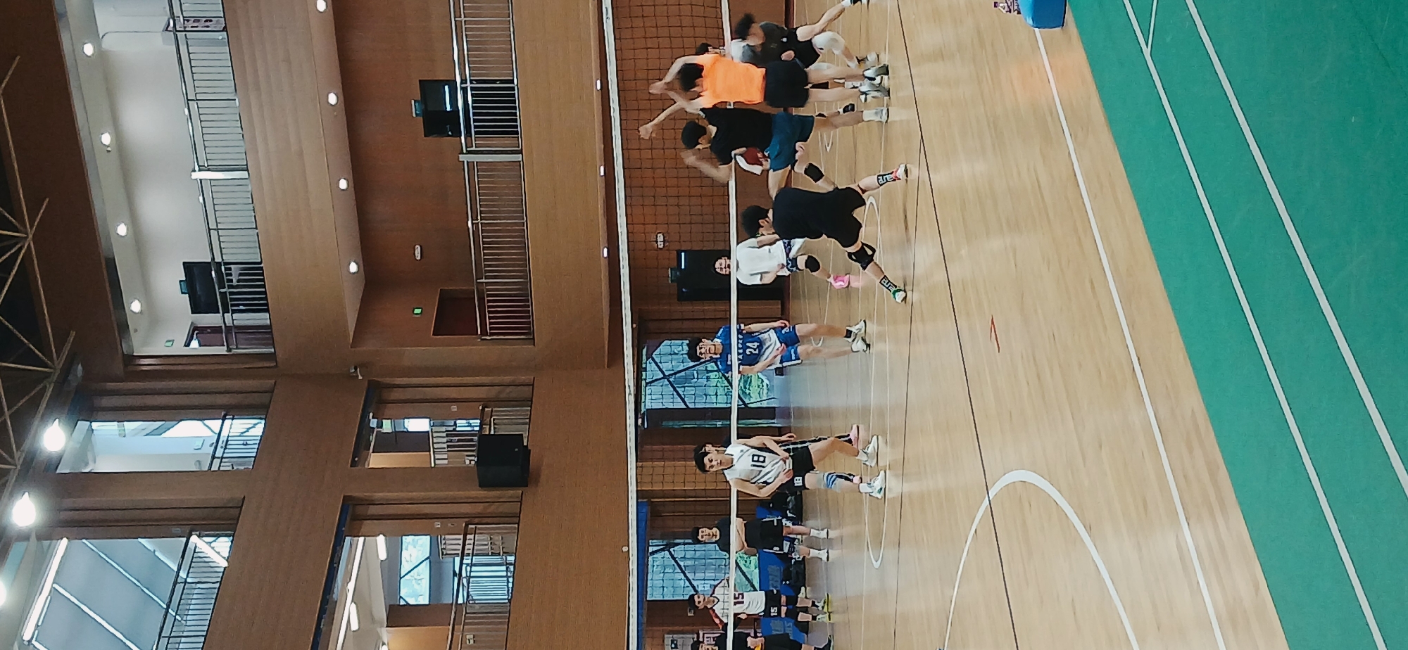 A volleyball game is in progress inside a gymnasium. There are two teams of players, one in blue and one in black/orange, positioned on either side of a net. Some players are jumping, indicating active play. The gymnasium has wooden floors with painted boundary lines. A green running track is visible along one side of the court. The walls are paneled with wood, featuring windows and a raised balcony area. The game is taking place in Chongqing High-tech Zone, China.