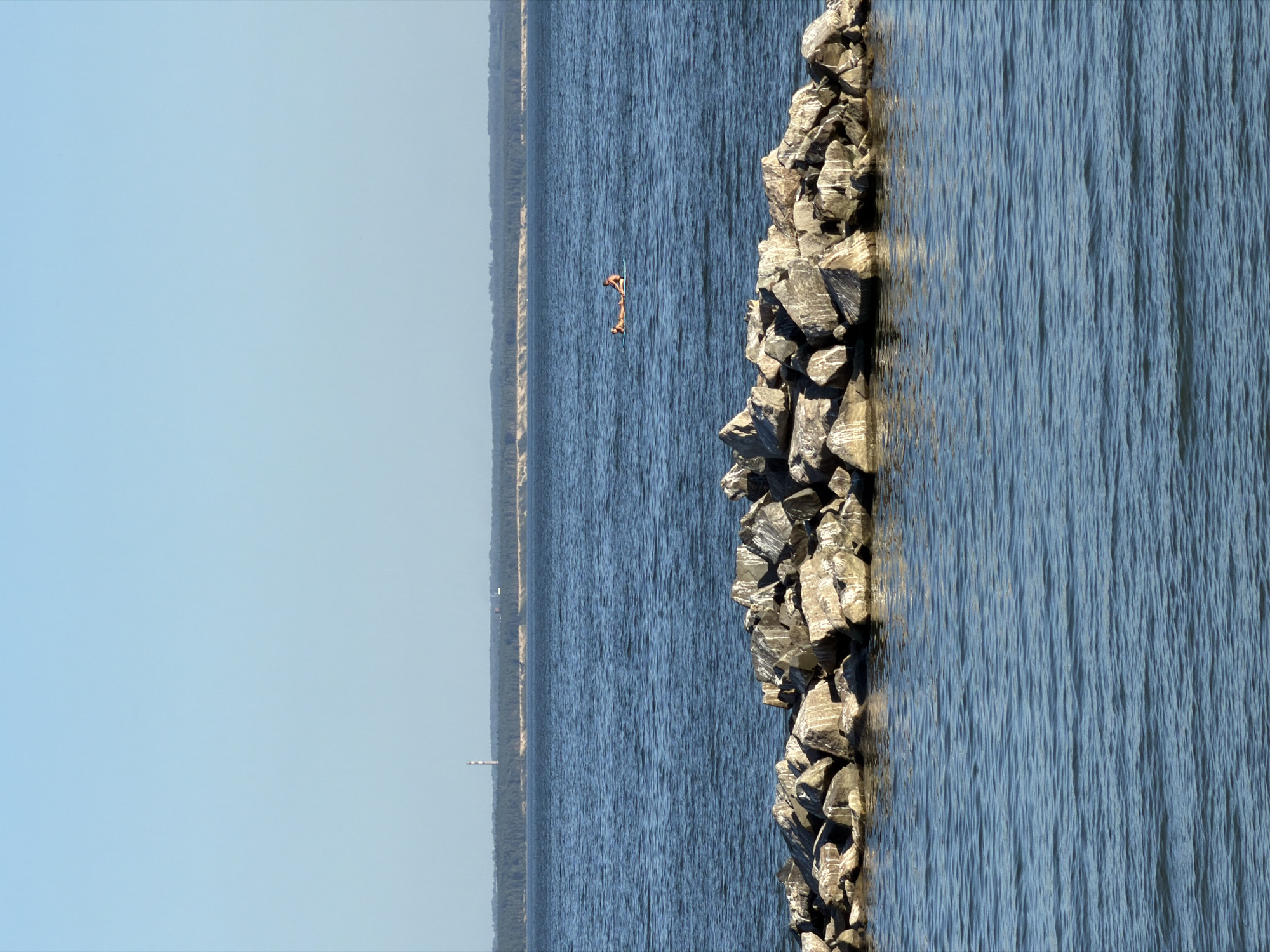 The image shows a body of water with a clear blue sky. In the foreground, there is a rocky breakwater. In the mid-ground, two people are lying on a flotation device in the water. In the background, a tree line is visible, and a tall, slender tower with a light at the top stands against the horizon. There is no indication that this scene is located in Novosibirsk, Russia; it is a generic outdoor scene.