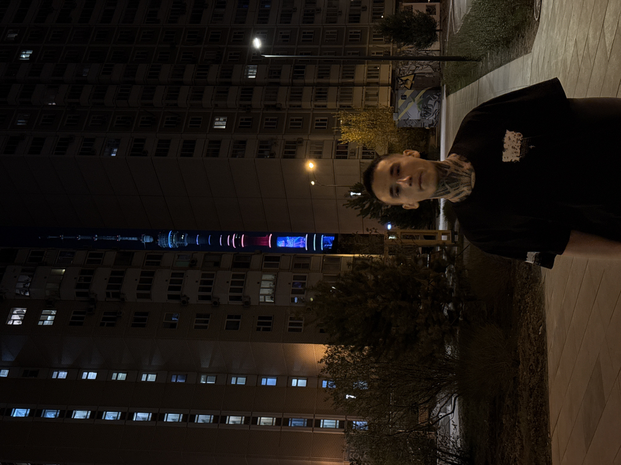 The image depicts a man standing on a sidewalk at night with a large building in the background, likely located in Khimki, Russia due to the presence of a tower illuminated with colored lights, similar to the Ostankino Tower but with a unique color configuration. The building has many illuminated windows. Streetlights are present, and there are some trees and vegetation visible. The man is wearing a dark t-shirt and has visible tattoos on his neck. The sidewalk appears to be paved.